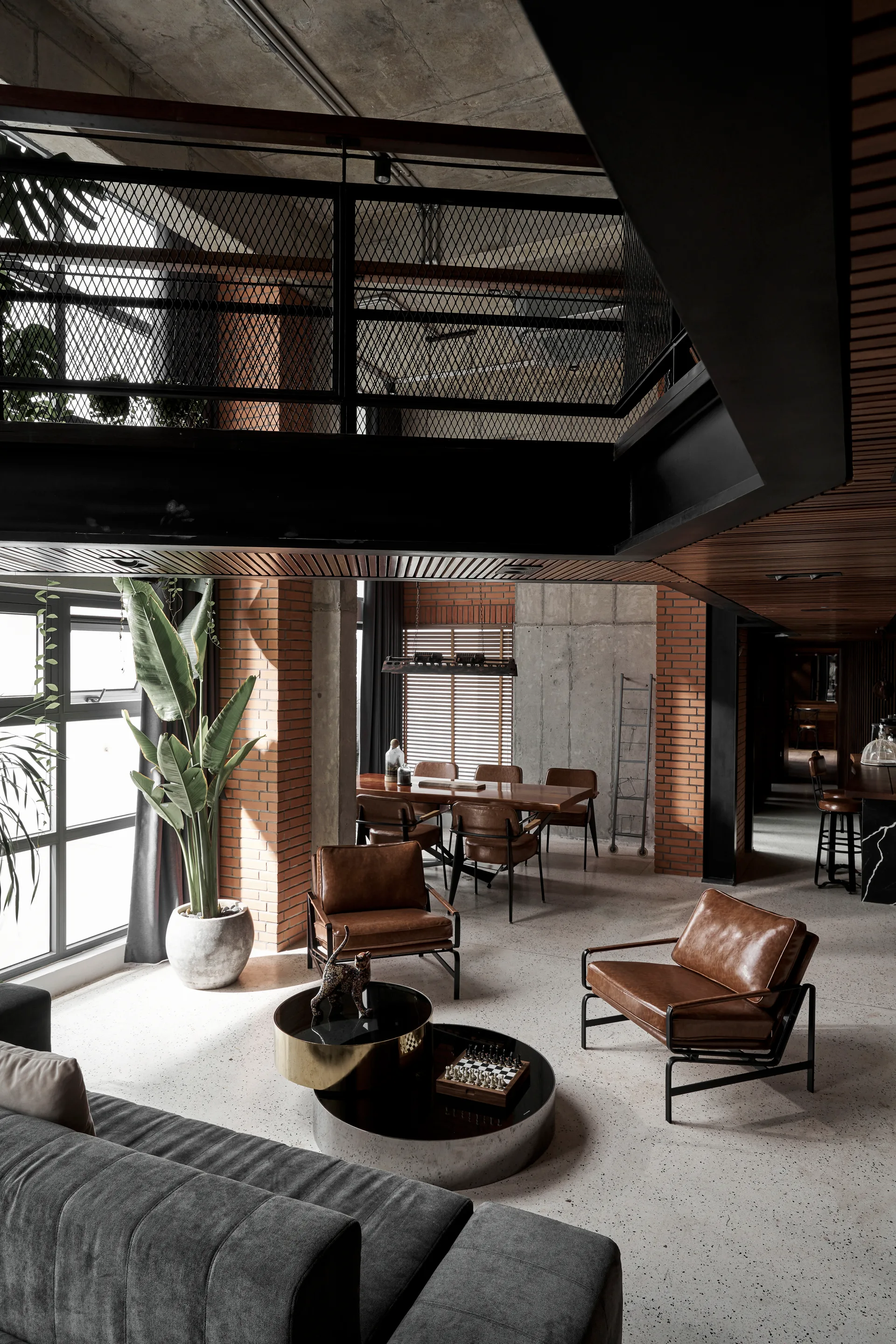 An industrial living room features brown leather armchairs, a grey sofa, exposed brick columns, and an upper mezzanine with black mesh railings.