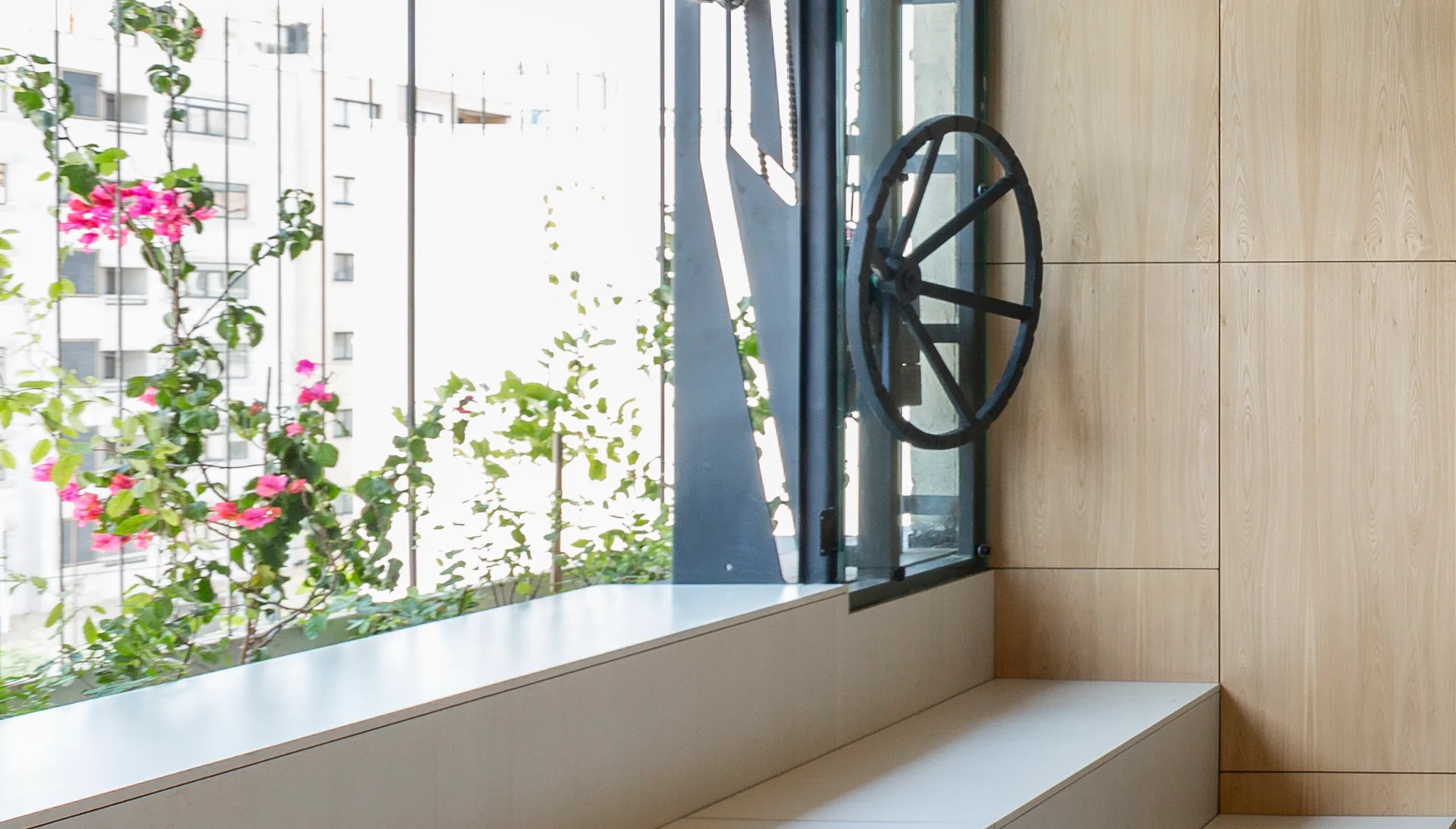 A large window with an industrial black wheel opener sits next to a pale wood paneled wall and a minimalist white bench.