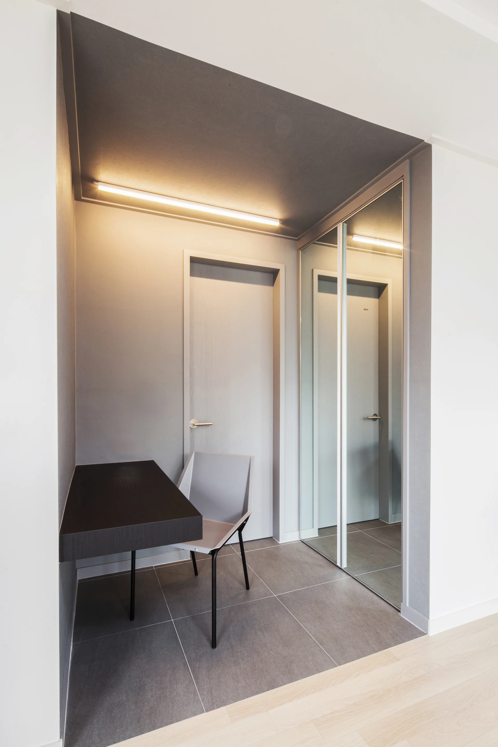 A minimalist gray entryway featuring a floating black desk, a light gray chair, and a large mirrored sliding closet door.
