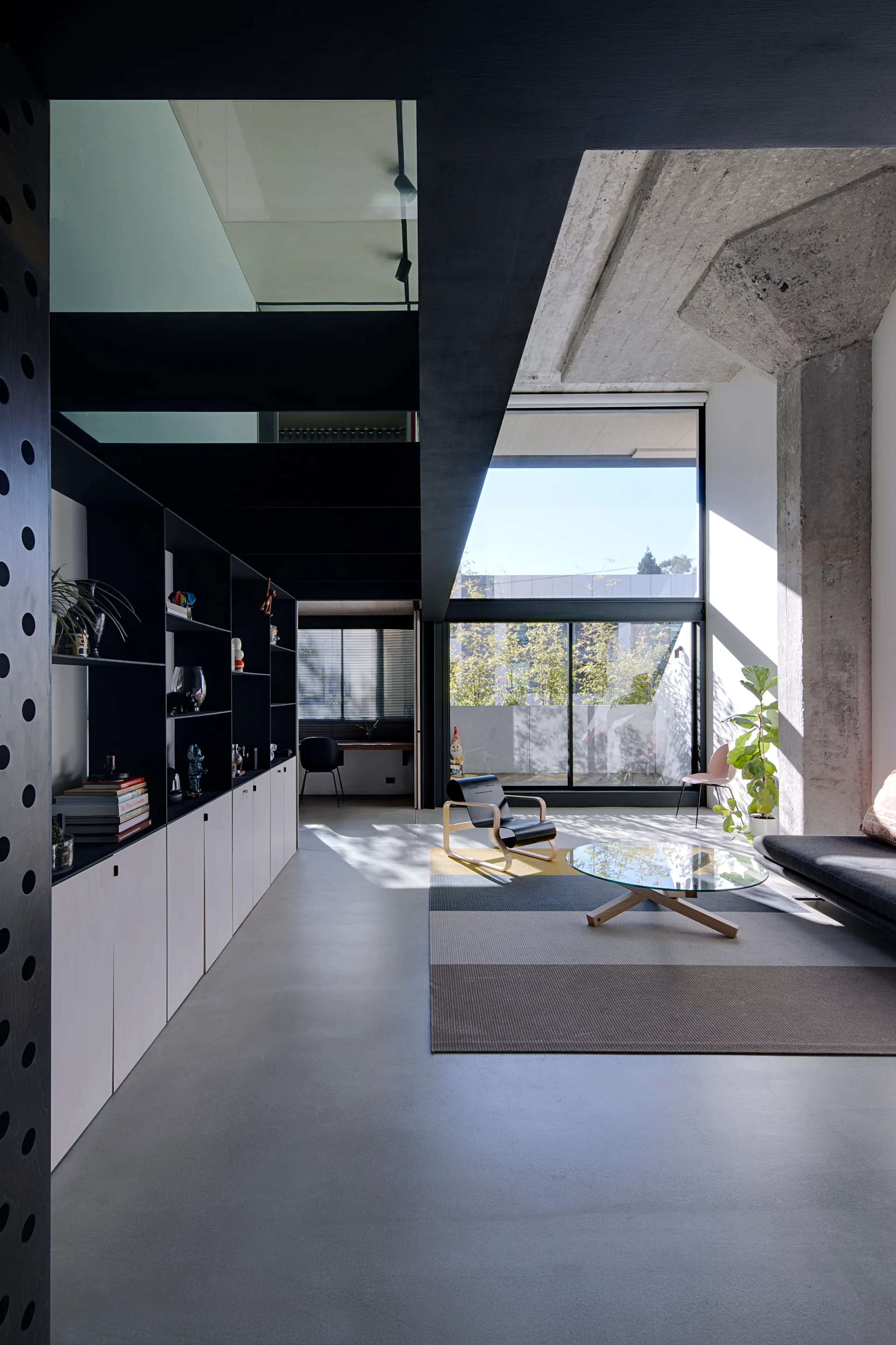 A modern double-height living room featuring a raw concrete column, custom dark shelving, a glass mezzanine floor panel, and a large window.