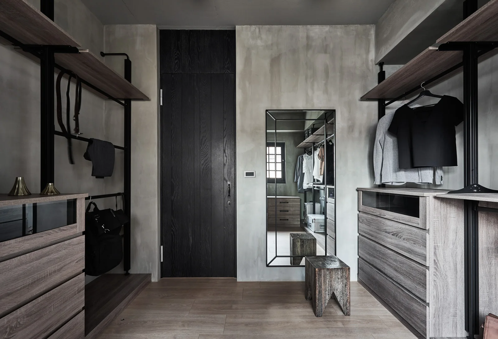 A modern walk-in closet features textured concrete-style walls, an open shelving system with drawers, a tall dark wood door, and a full-length mirror.