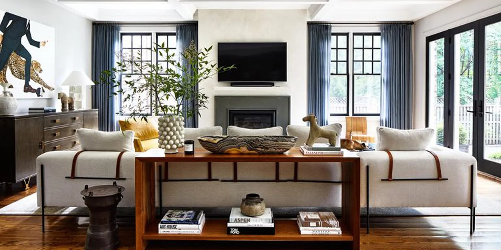 A symmetrical living room featuring two white sofas with leather back straps, a wooden console table, a central fireplace, and blue drapes.