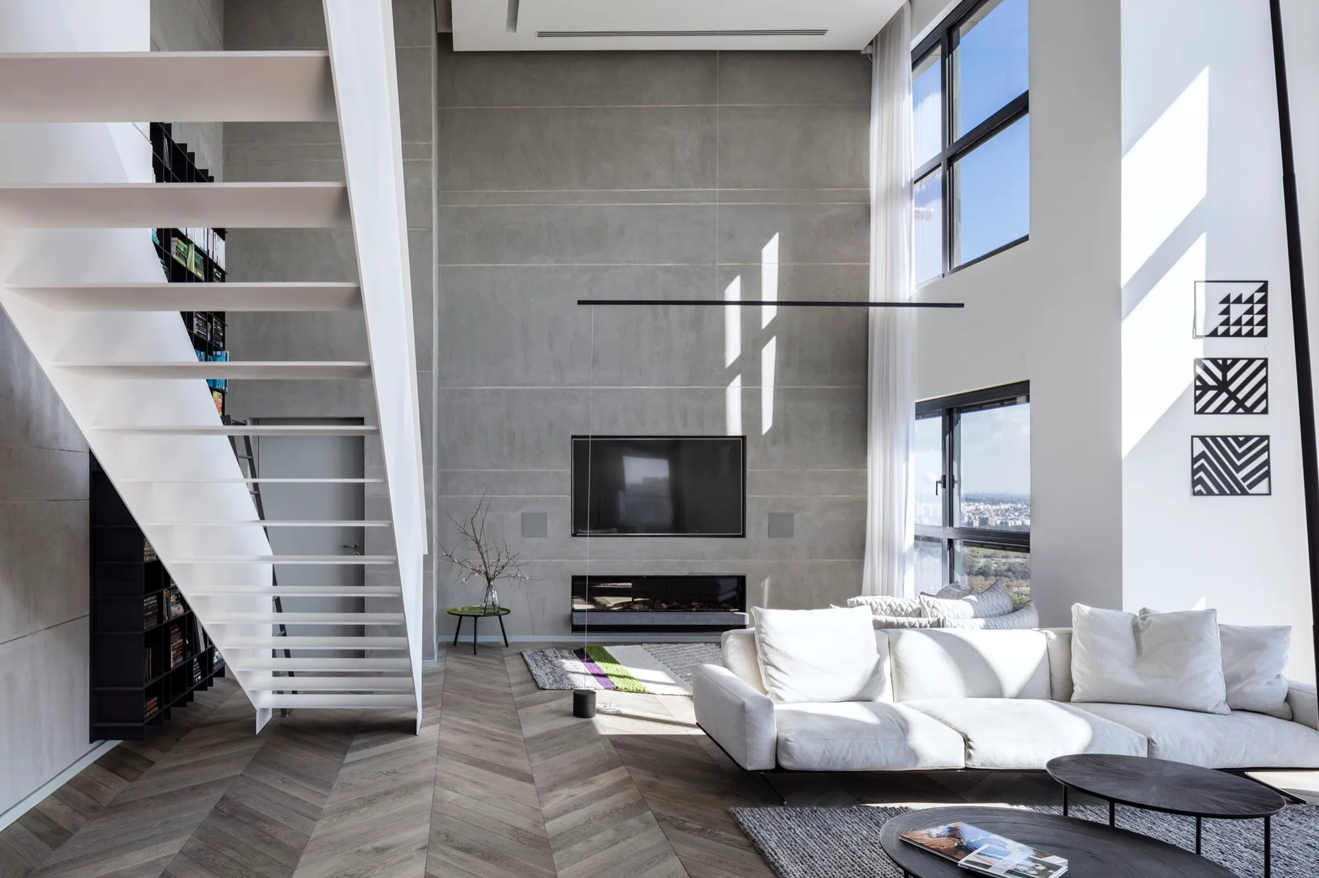 A spacious double-height living room featuring a white floating staircase, a grey paneled fireplace wall, a white sectional sofa, and herringbone floors.