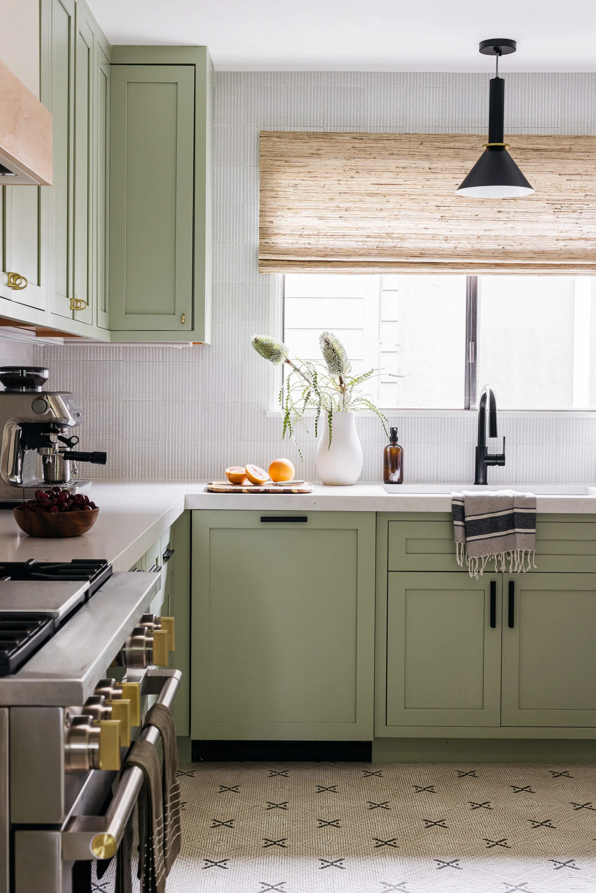 A bright kitchen features sage green shaker cabinets, a subtle tile backsplash, a black gooseneck faucet, and a woven natural window shade.