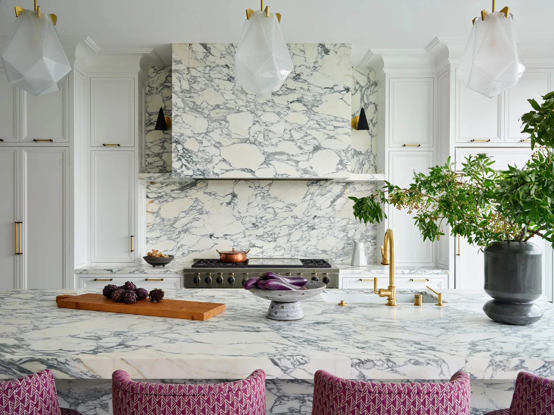 A bright kitchen features a large marble island, a matching marble-clad range hood, white cabinets, and patterned pink bar stools in the foreground.