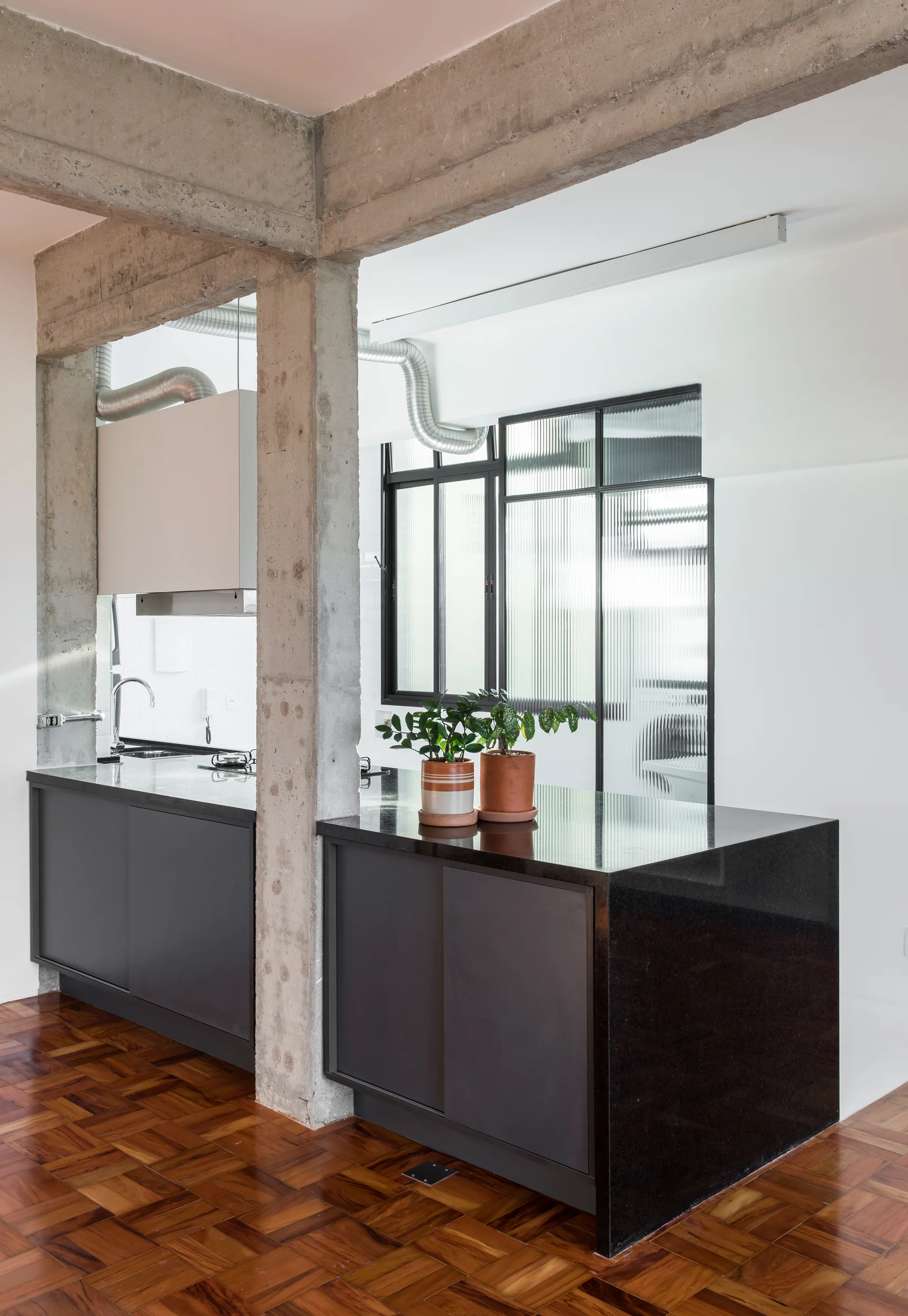 A modern kitchen island built around an exposed concrete column features black countertops, matte gray cabinets, and a fluted glass partition.