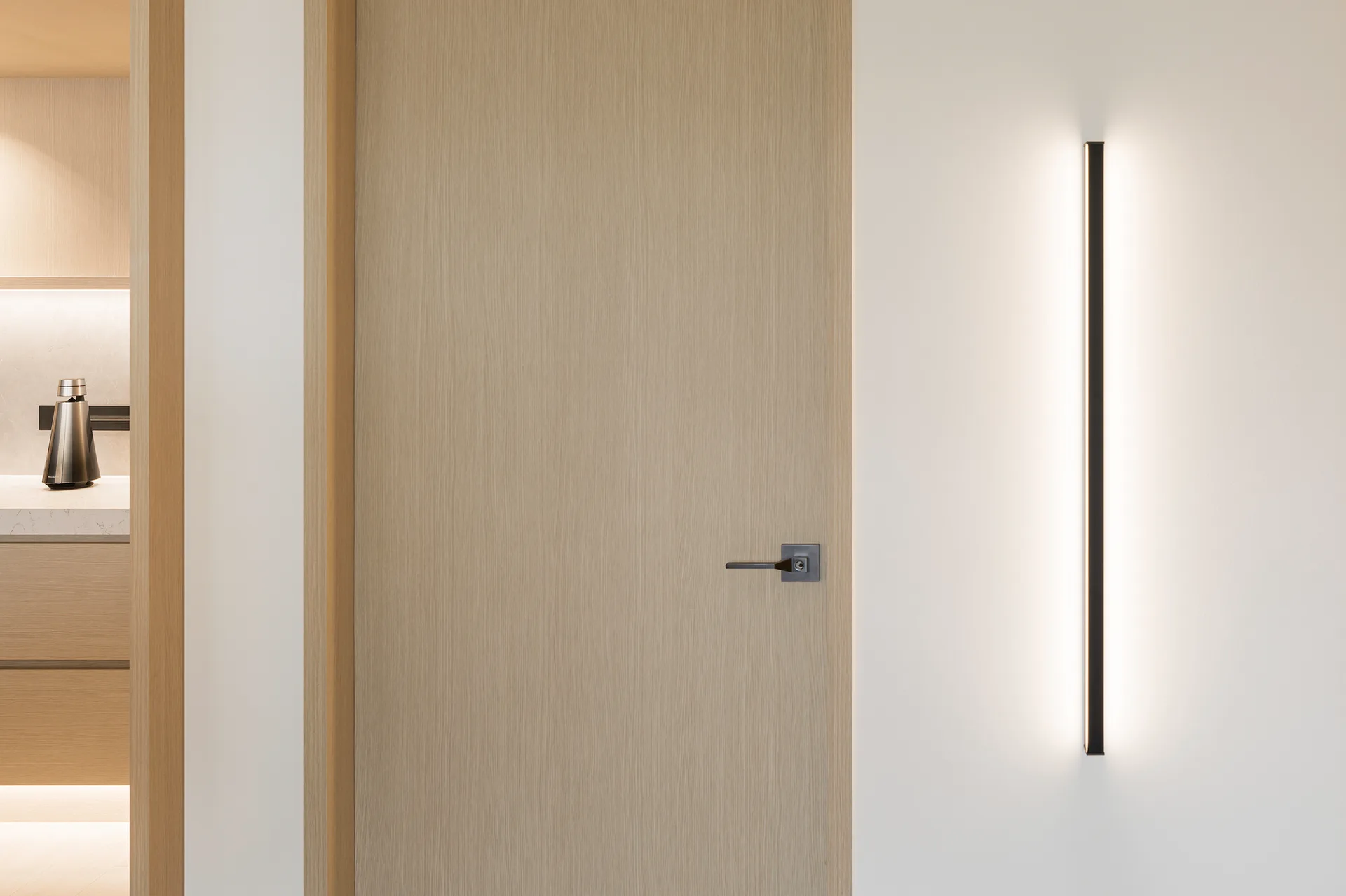 A light wood door with a dark metal handle stands next to a vertical linear sconce emitting a warm glow on a white wall.