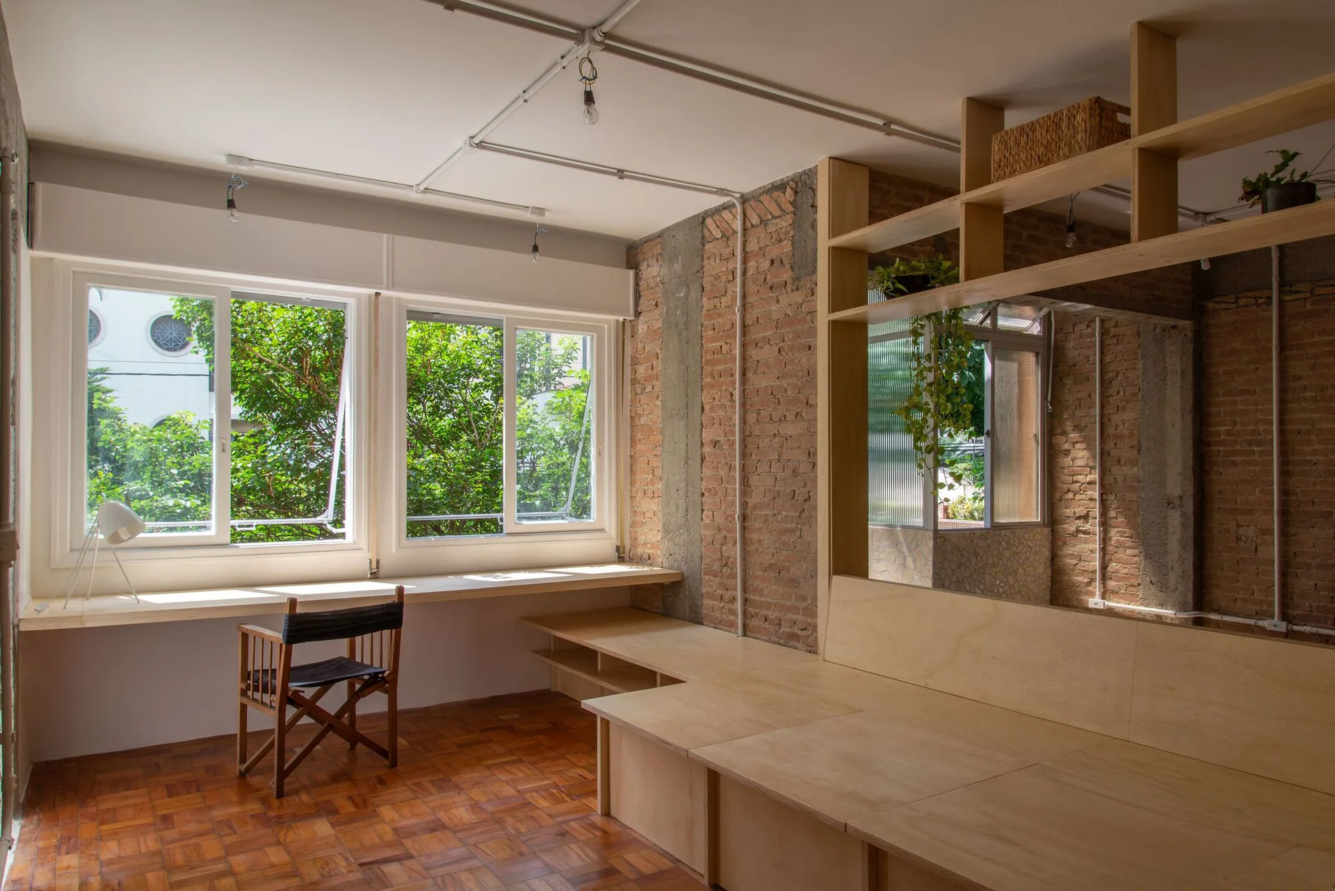A home office features a long wooden desk by a window, a wooden platform with shelves, parquet flooring, and an exposed brick wall.