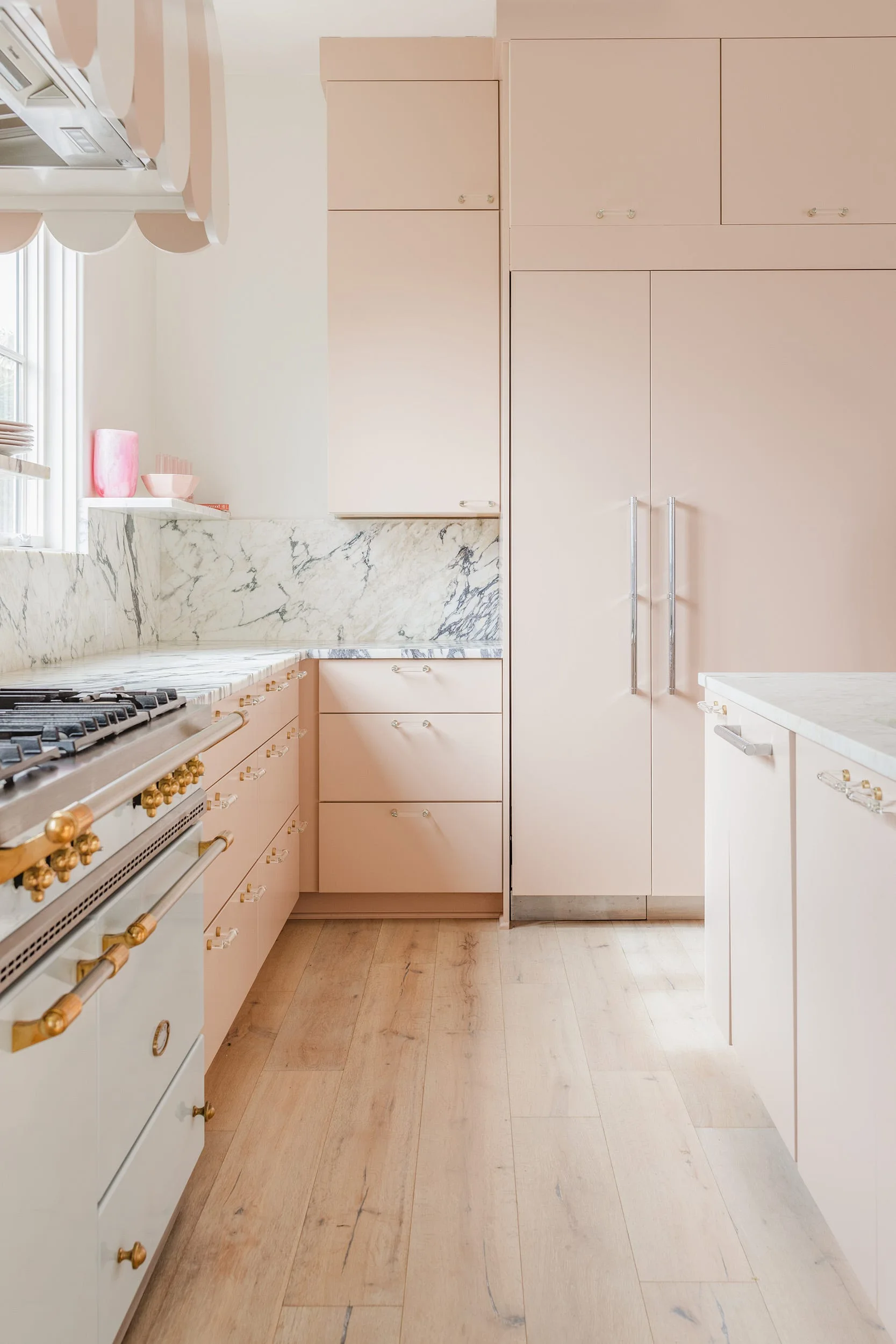 A residential kitchen features blush pink flat-panel cabinets, a white veined marble backsplash and countertops, and a white range stove.