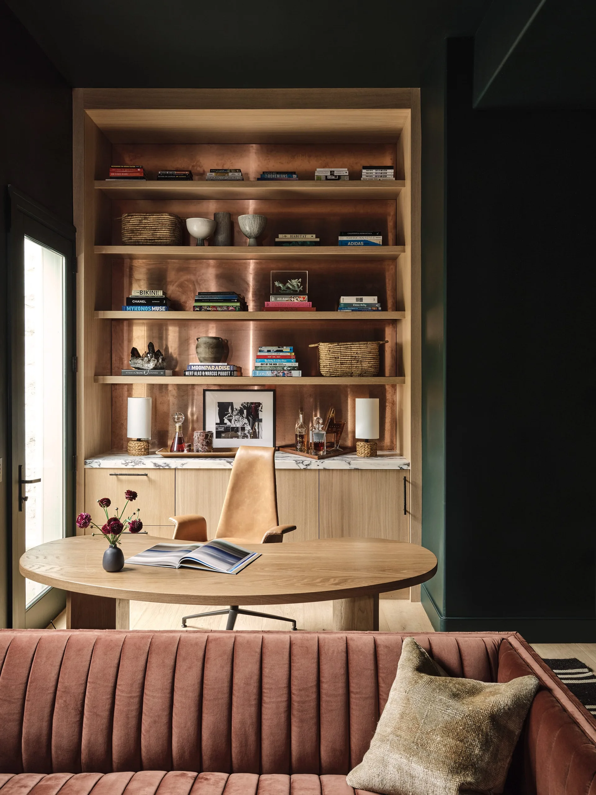 A home office features a wooden oval desk, a leather swivel chair, and custom wood shelving with a reflective copper backing panel.