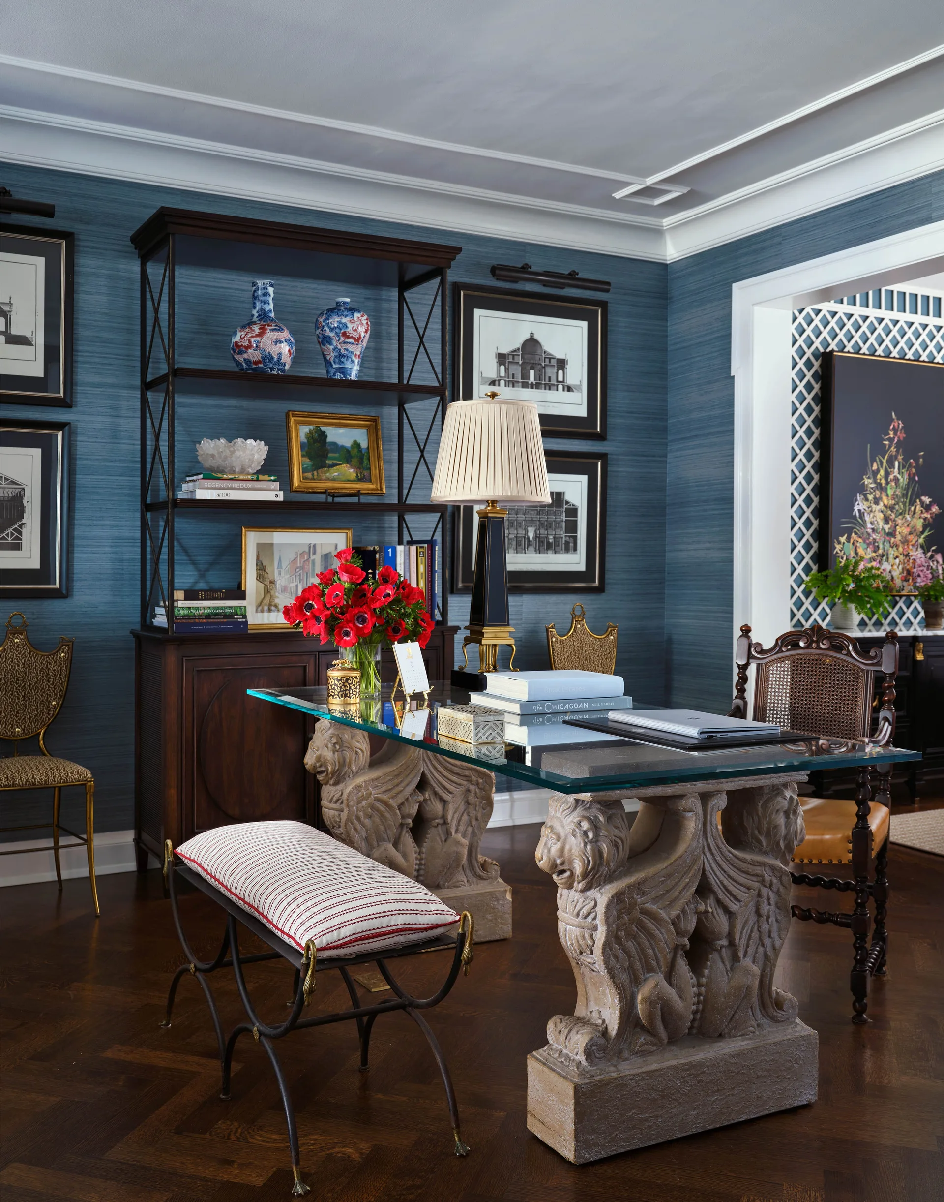 A classic home office featuring a glass desk with stone lion bases, textured blue wallpaper, a dark wood bookcase, and traditional artwork.
