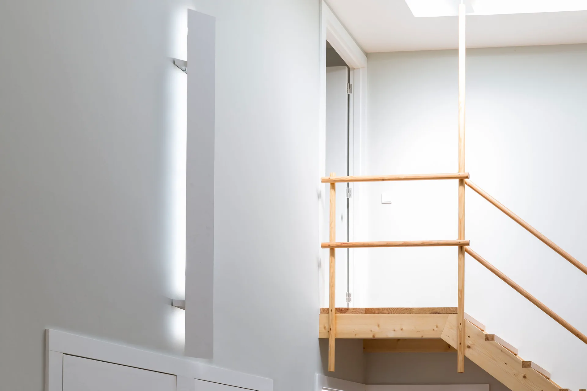 A minimalist white staircase features a light wood handrail and a tall vertical wall-mounted light fixture.