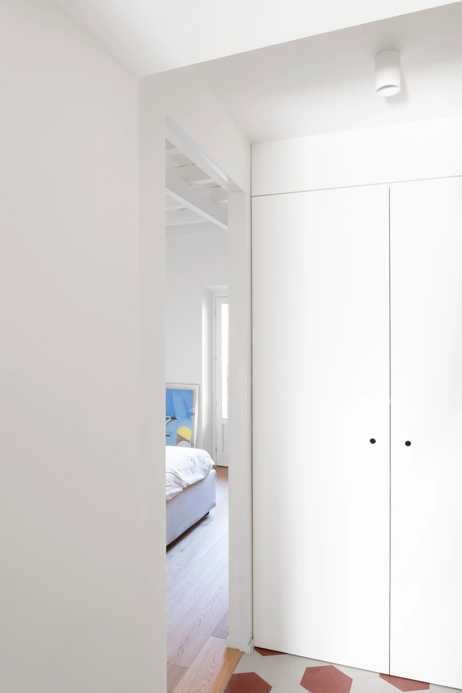 A view from a white hallway with minimalist built-in closet doors and hexagonal floor tiles looking into a bedroom with light wood flooring.