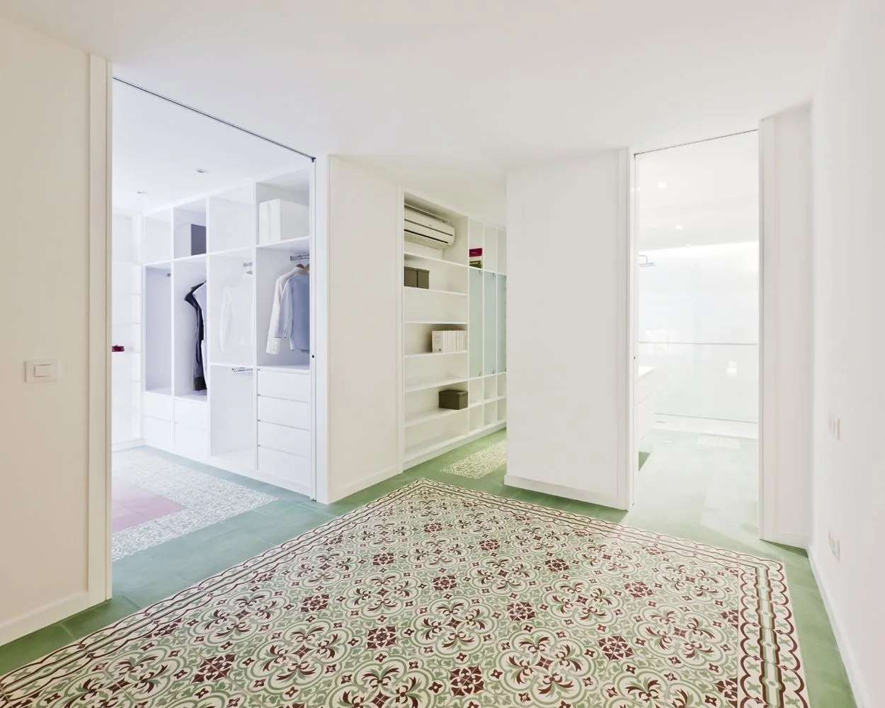 A bright white dressing room features custom built-in shelving, open doorways, and green and maroon patterned ceramic tile flooring.