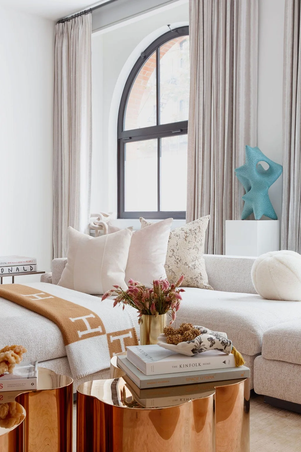 A light-filled living room featuring a cream boucle sectional sofa, metallic gold cylinder coffee tables, and an arched black-framed window.