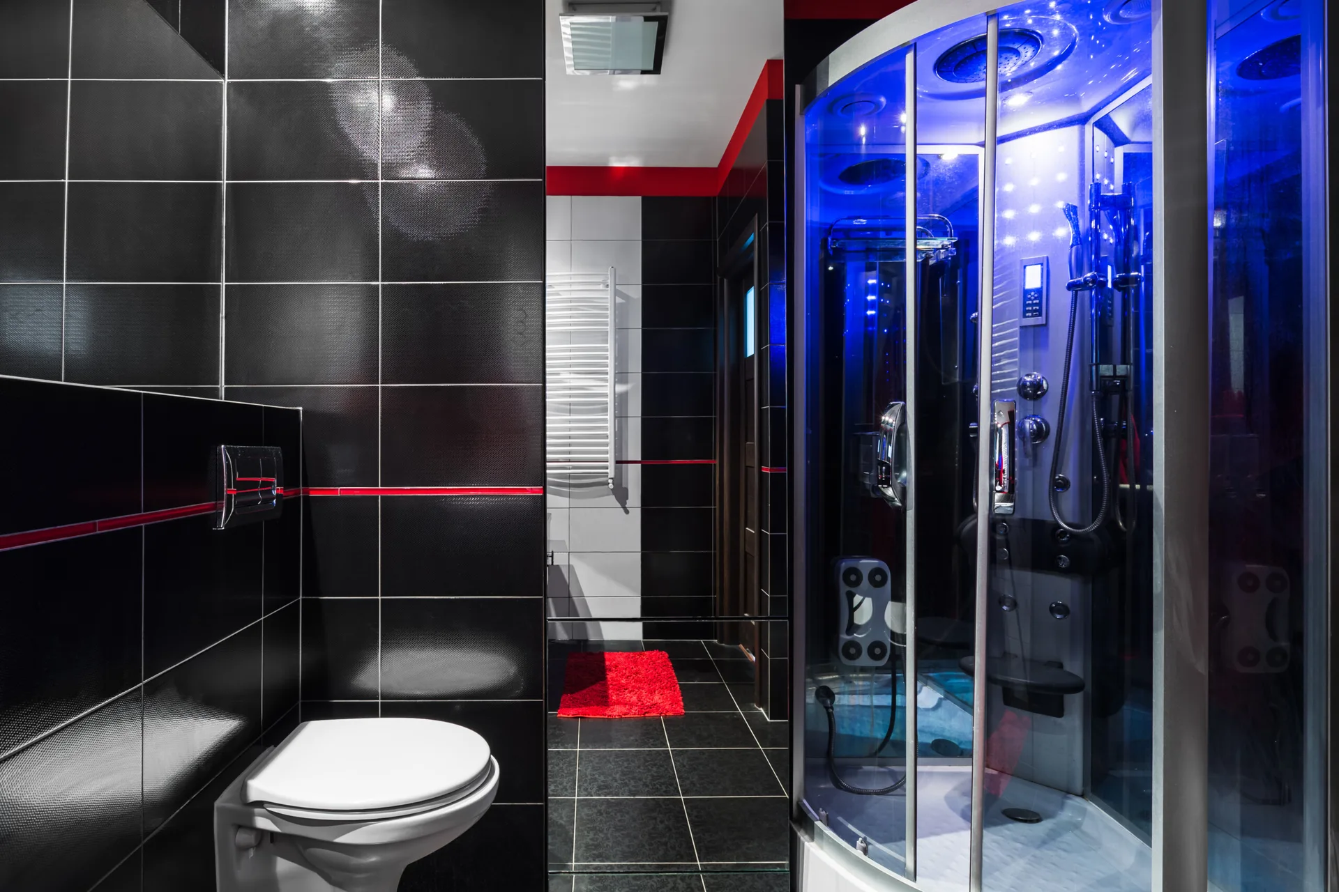 A modern bathroom featuring glossy black wall tiles, a wall-mounted toilet, and a glass shower enclosure illuminated with a vibrant blue glow.