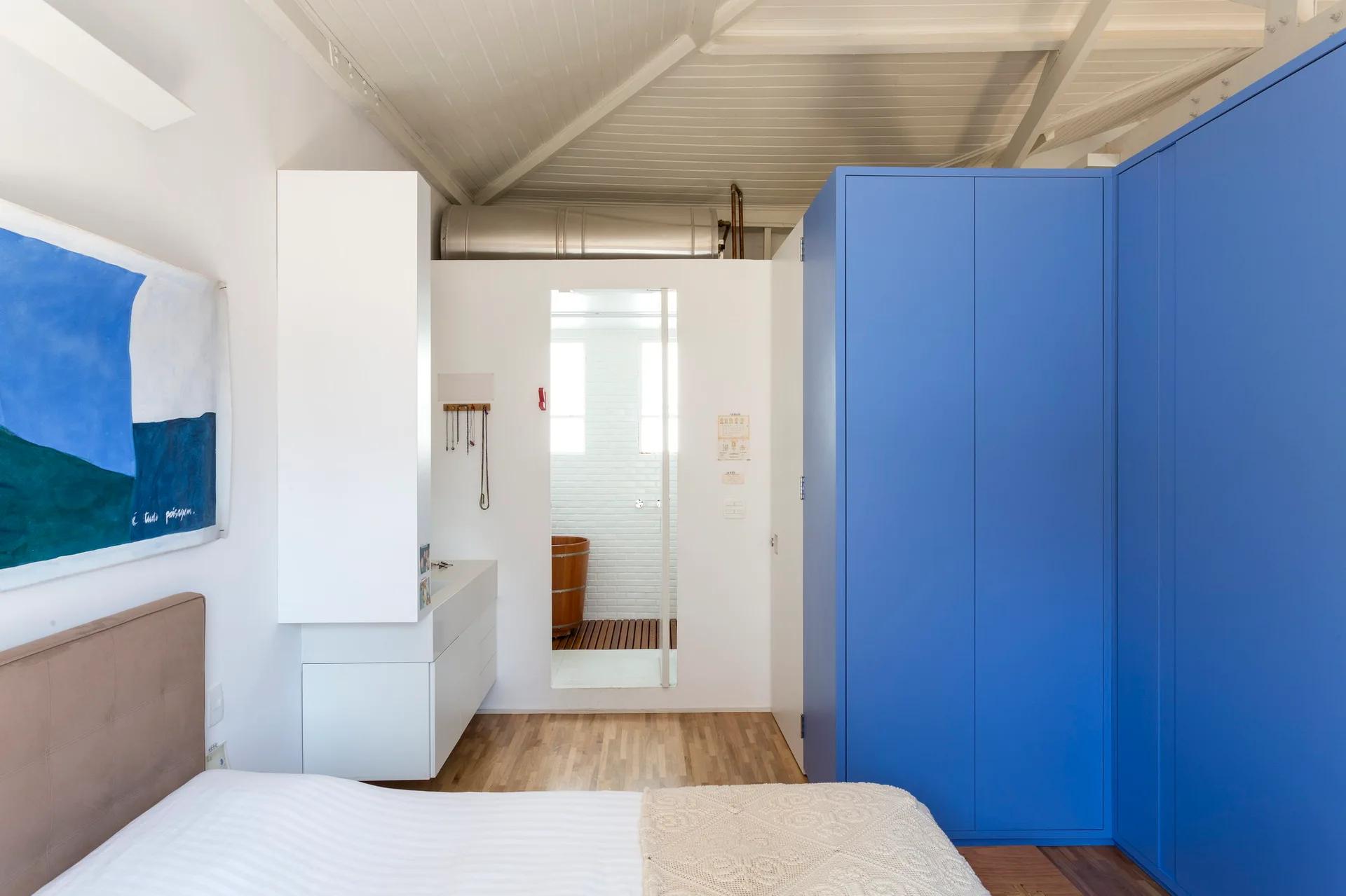 A modern bedroom with a large blue wardrobe, a bed, and a view into a bathroom with a wooden soaking tub.