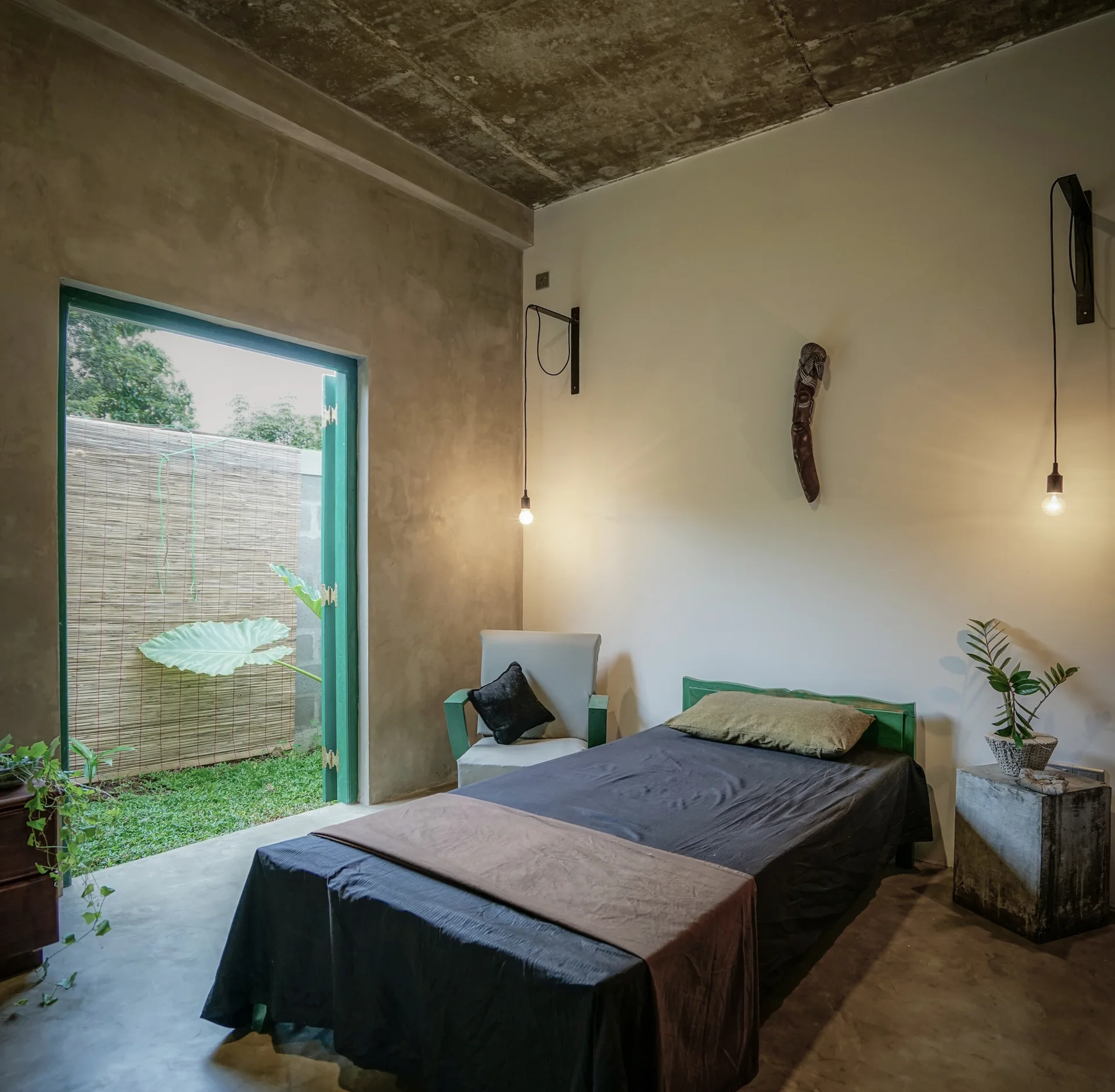 A minimalist bedroom with a single bed, raw concrete walls and ceiling, bare bulb wall pendant lights, and an open doorway showing a green garden.