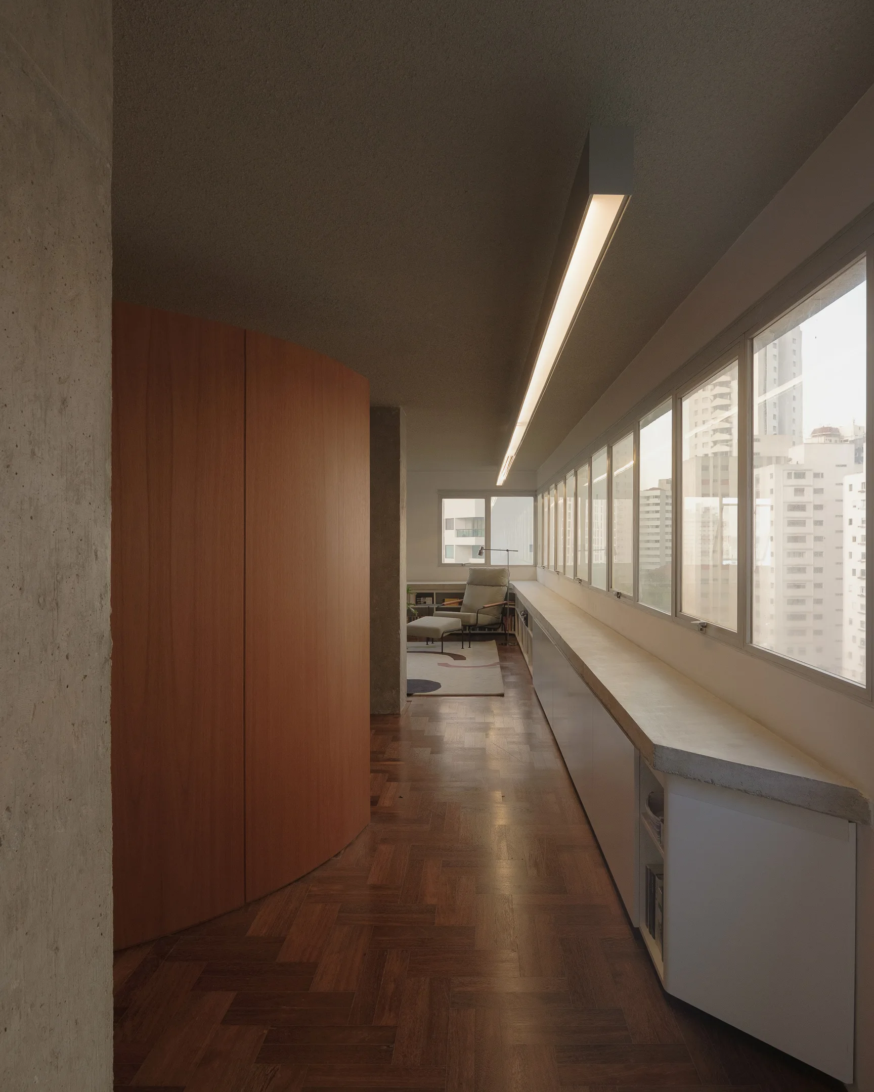 A minimalist hallway corridor featuring a curved natural wood partition wall, herringbone patterned flooring, and built-in white cabinets under wide windows.