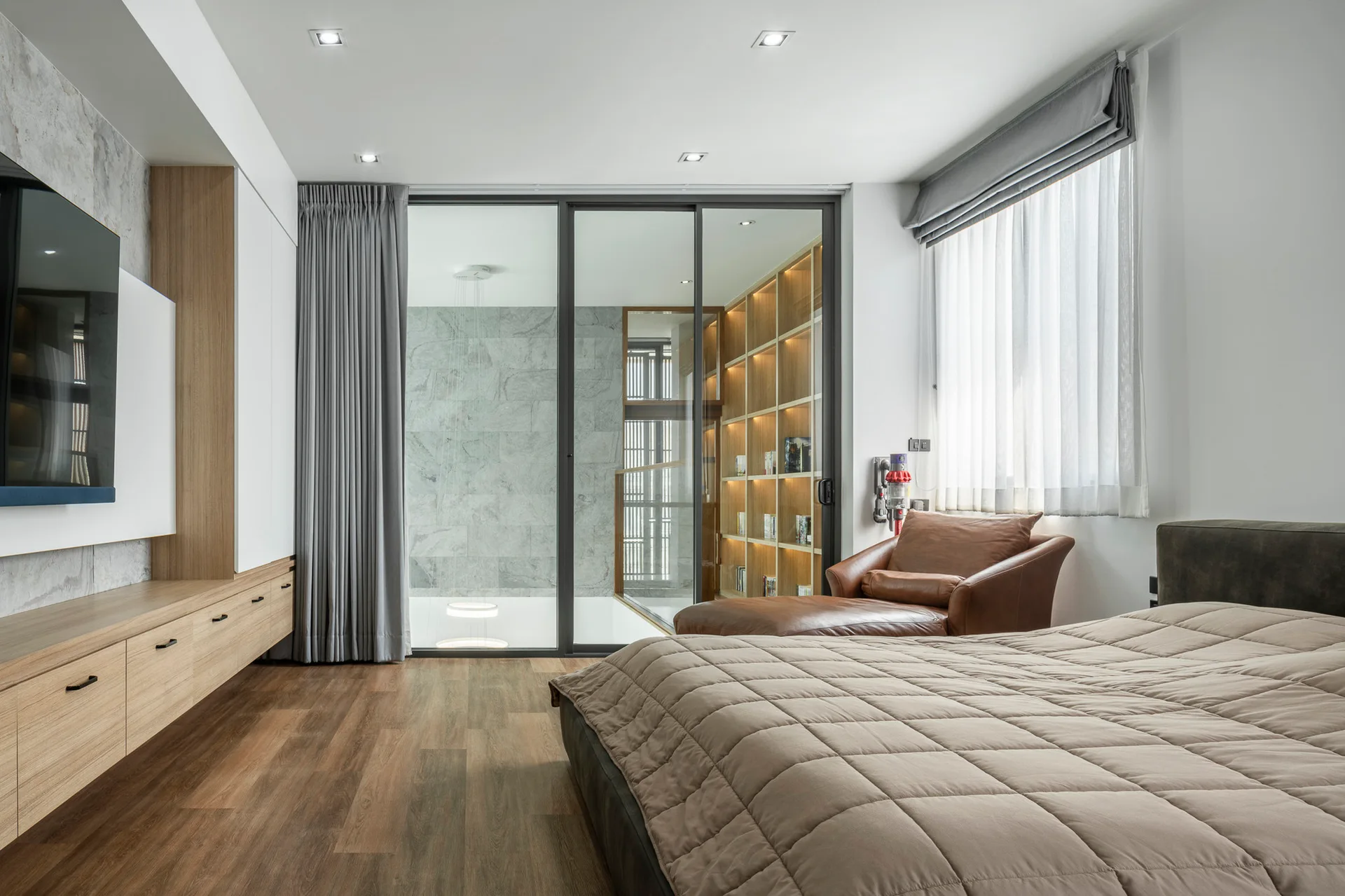 A minimalist bedroom features a quilted bedspread, a brown leather lounge chair, and a large sliding glass partition looking into an illuminated hallway.