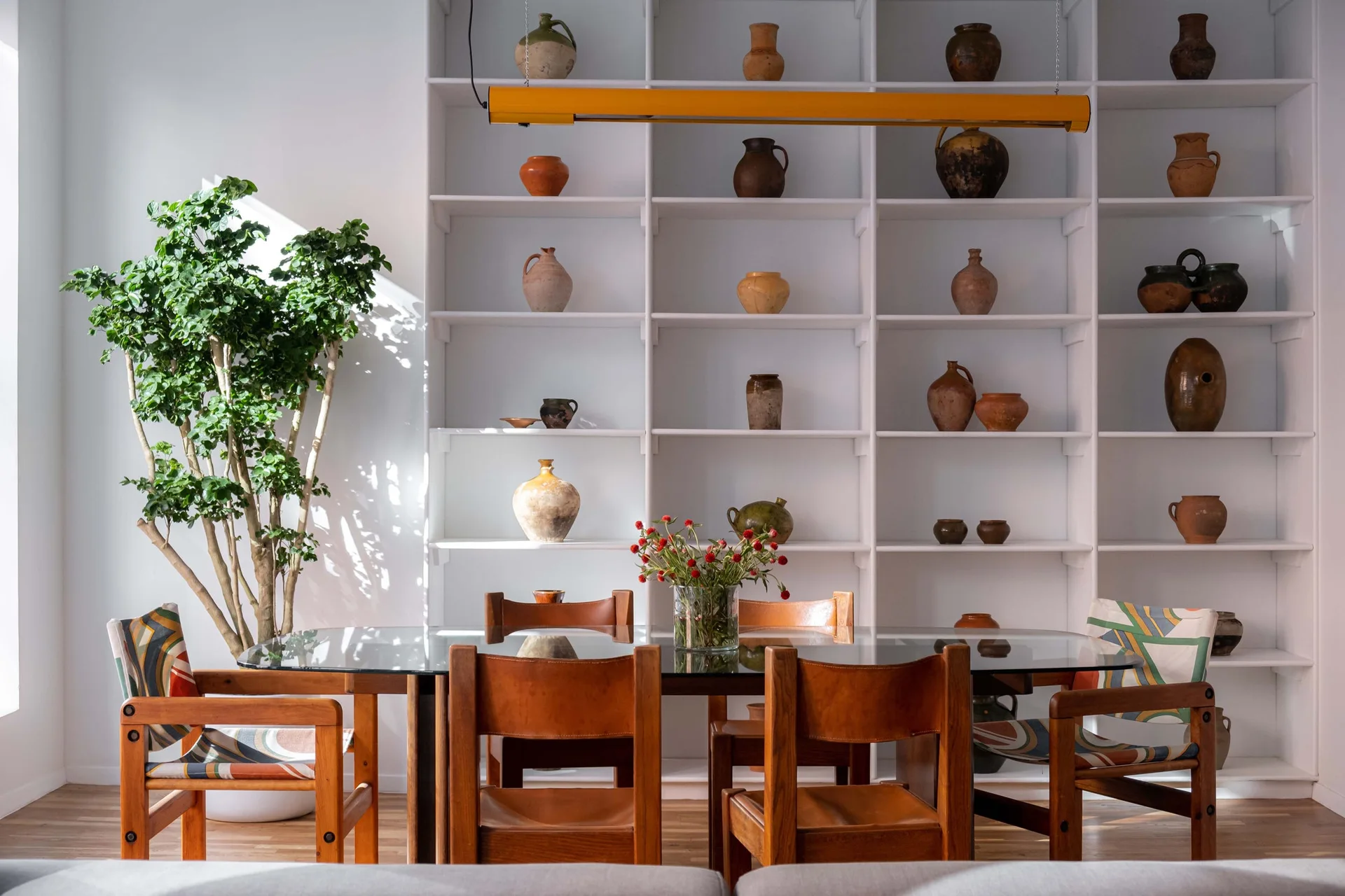 A glass dining table surrounded by wooden chairs sits in front of large white shelving filled with rustic ceramic pottery.