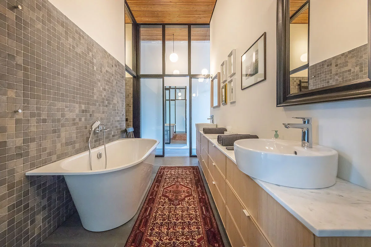 A narrow bathroom features a freestanding bathtub, a gray mosaic tile wall, a long light wood vanity, and a patterned runner rug on the floor.