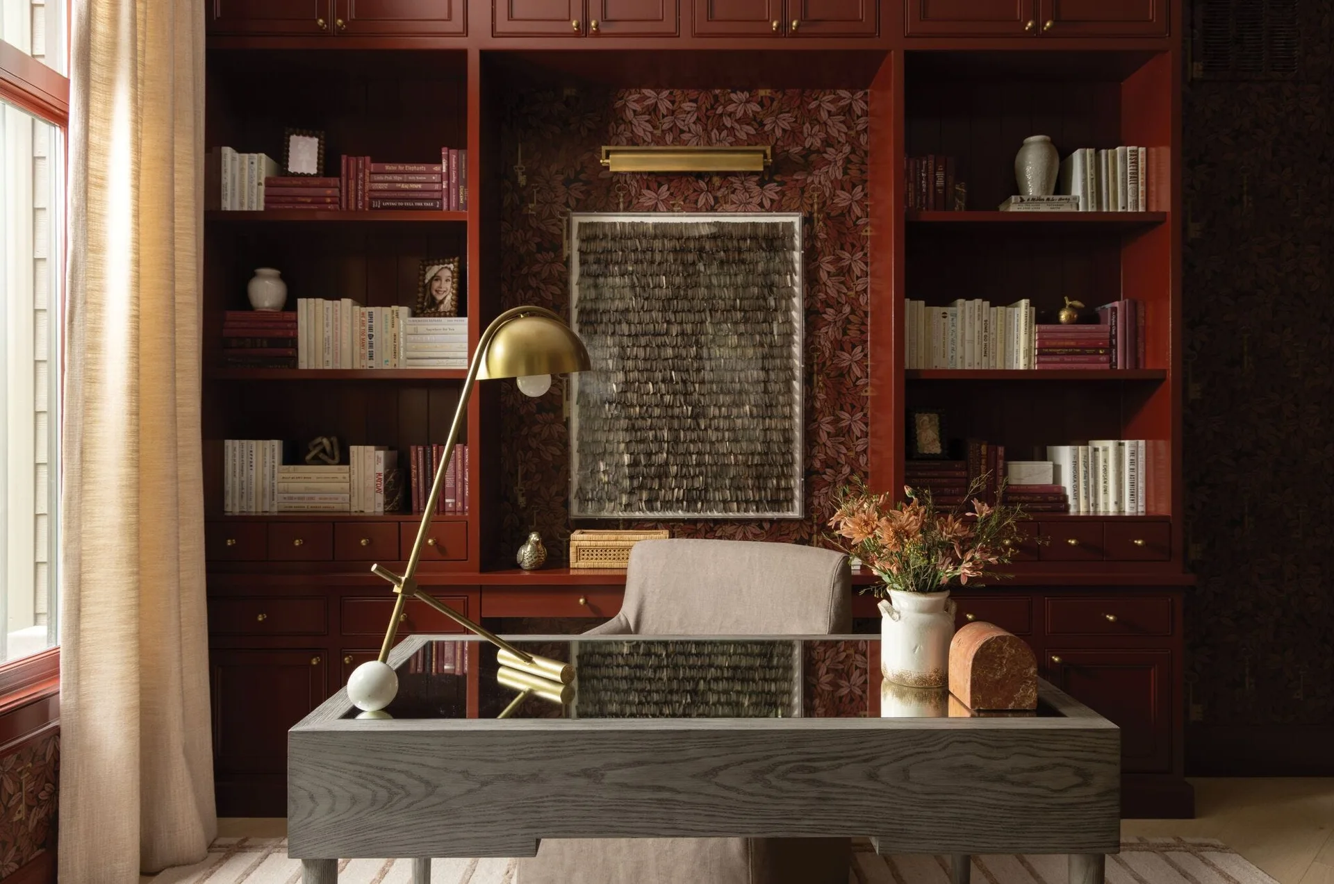 A grey wooden desk with a mirrored top sits before a wall of dark red built-in bookshelves featuring floral wallpaper and brass lighting.