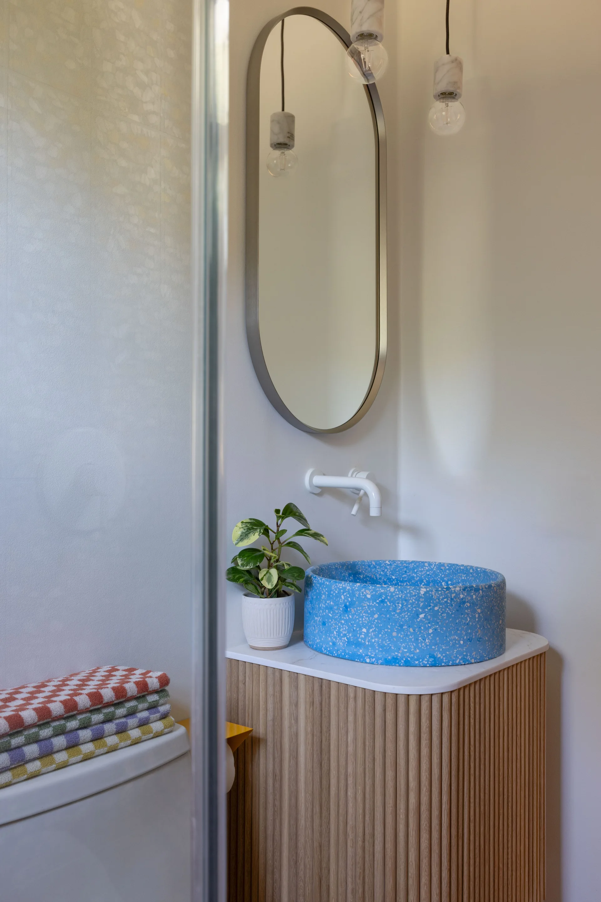 A modern bathroom vanity with a fluted wood base, a bright blue speckled terrazzo vessel sink, a white wall-mounted faucet, and an oval mirror.