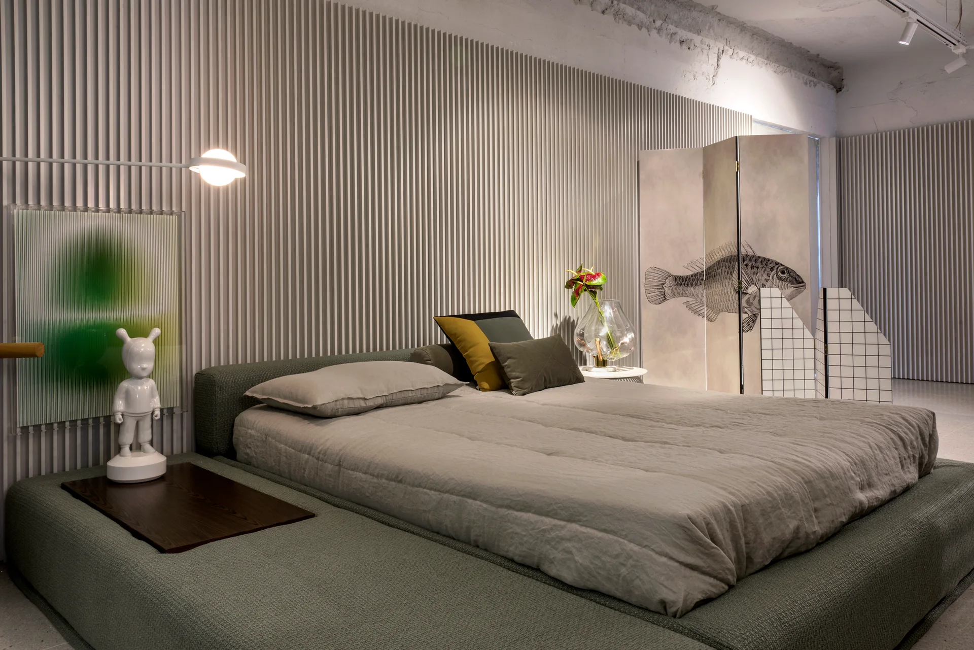 A low-profile grey upholstered bed sits against a white fluted panel wall featuring a modern sphere light and a decorative folding screen.