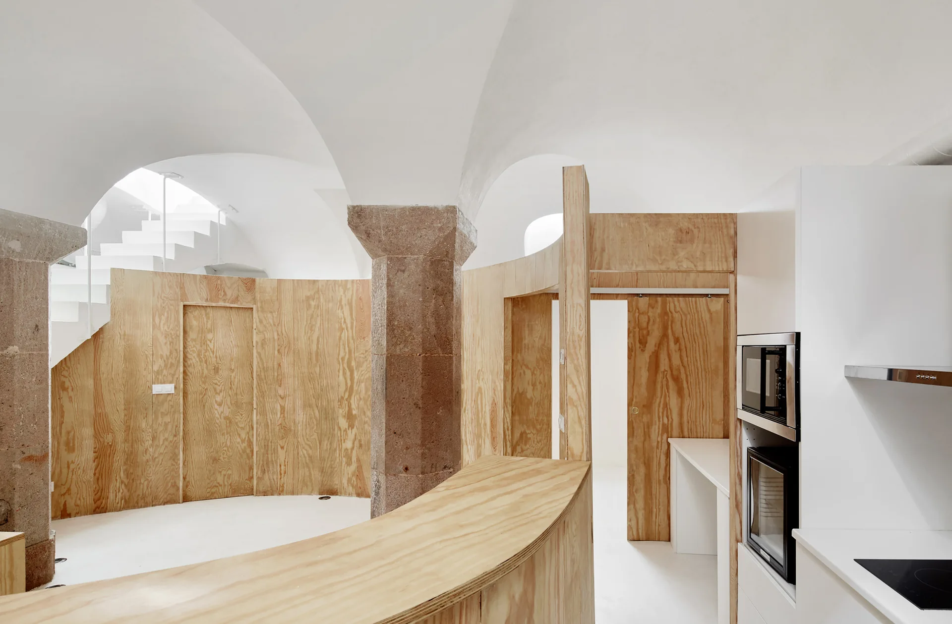 A modern minimalist kitchen featuring curved plywood partition walls, a central stone pillar, and white vaulted ceilings.