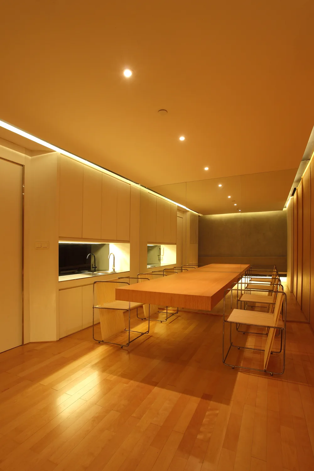A long wood dining table and metal-framed chairs sit on wood floors in a minimalist room with warm cove lighting and a mirrored wall.