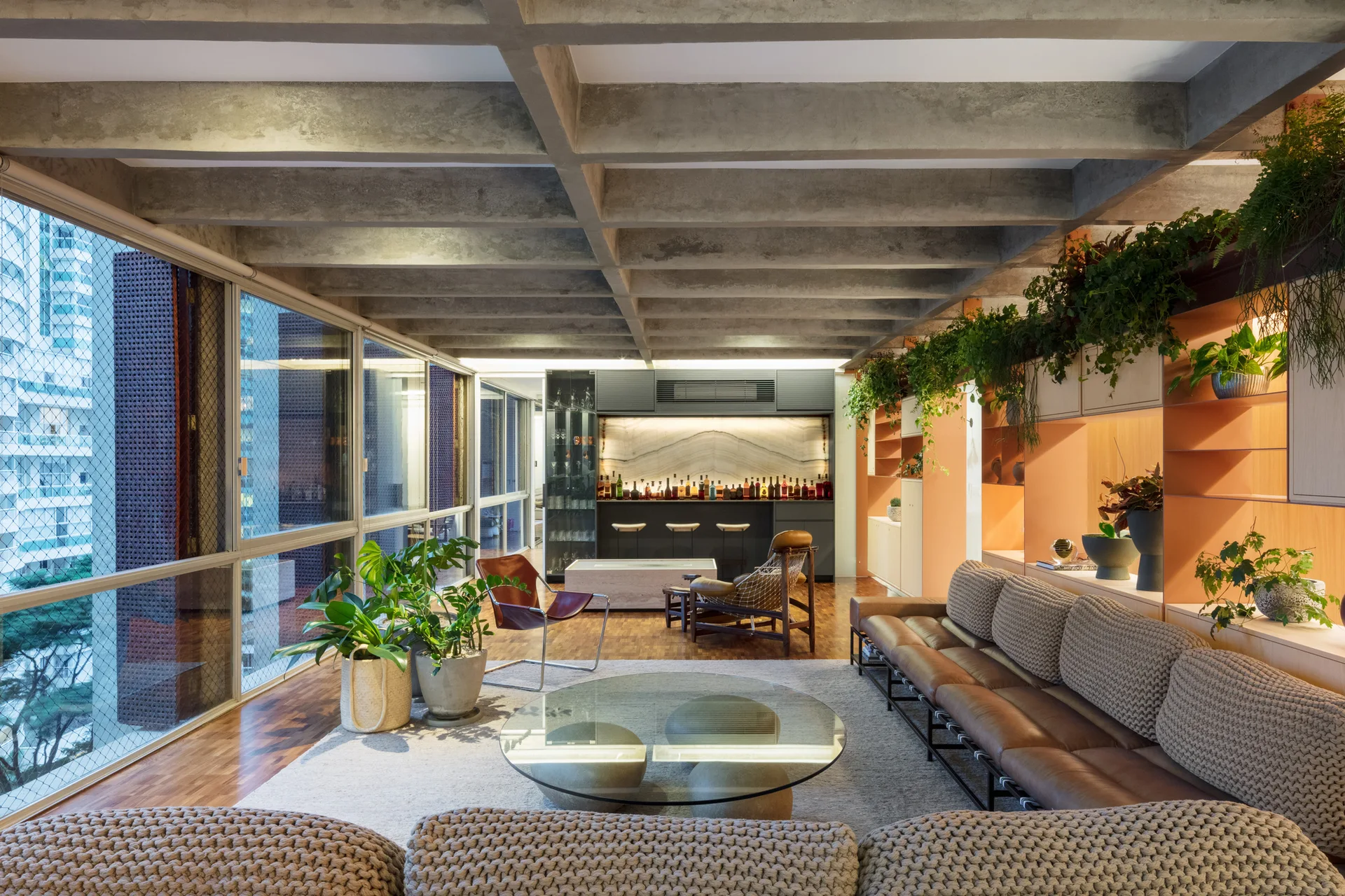 A modern living room features an exposed concrete grid ceiling, a long leather sofa, and an orange built-in shelving unit filled with potted plants.