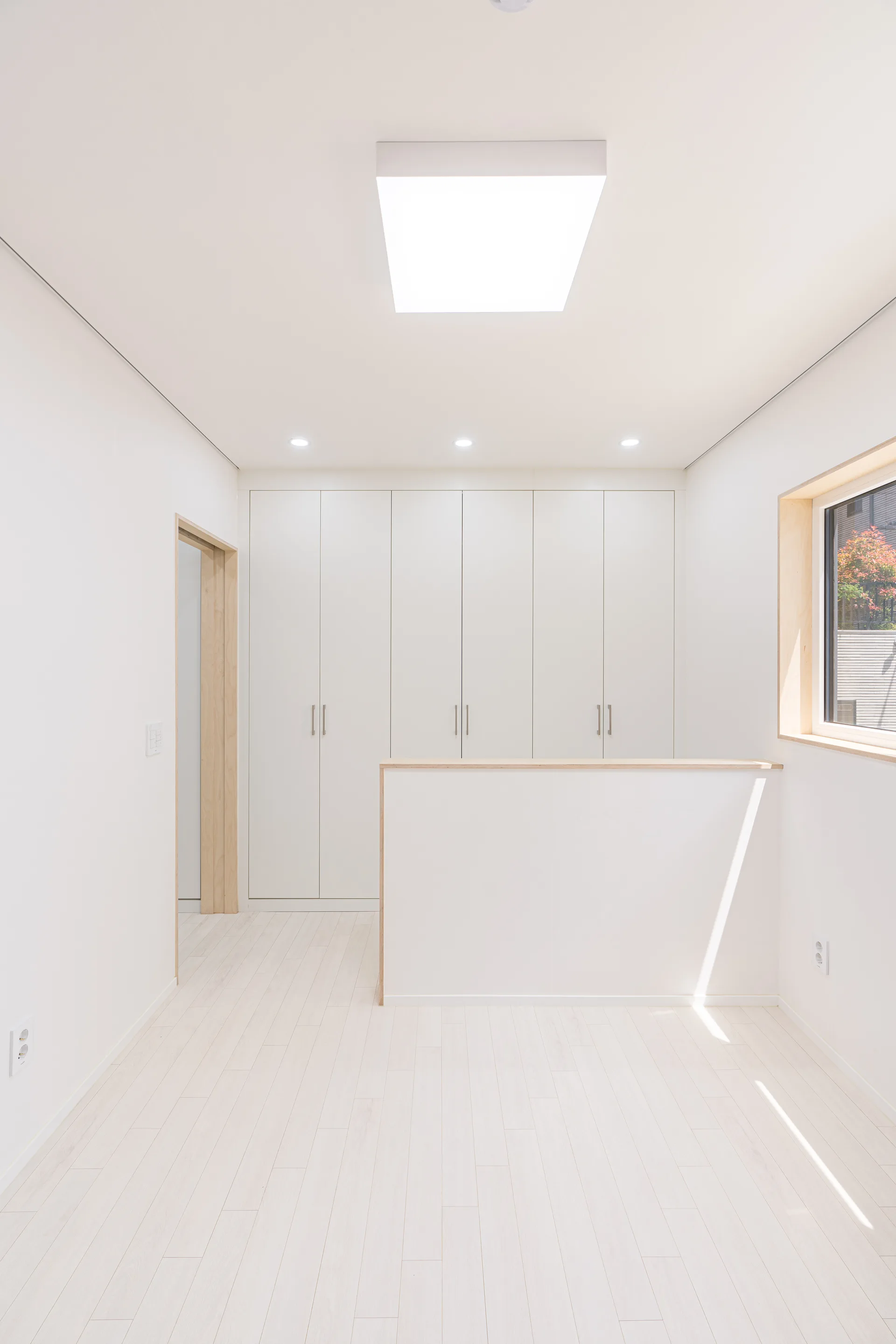 A minimalist white landing features built-in cabinets, a low partition wall with light wood trim, wood doorway frames, and light plank flooring.