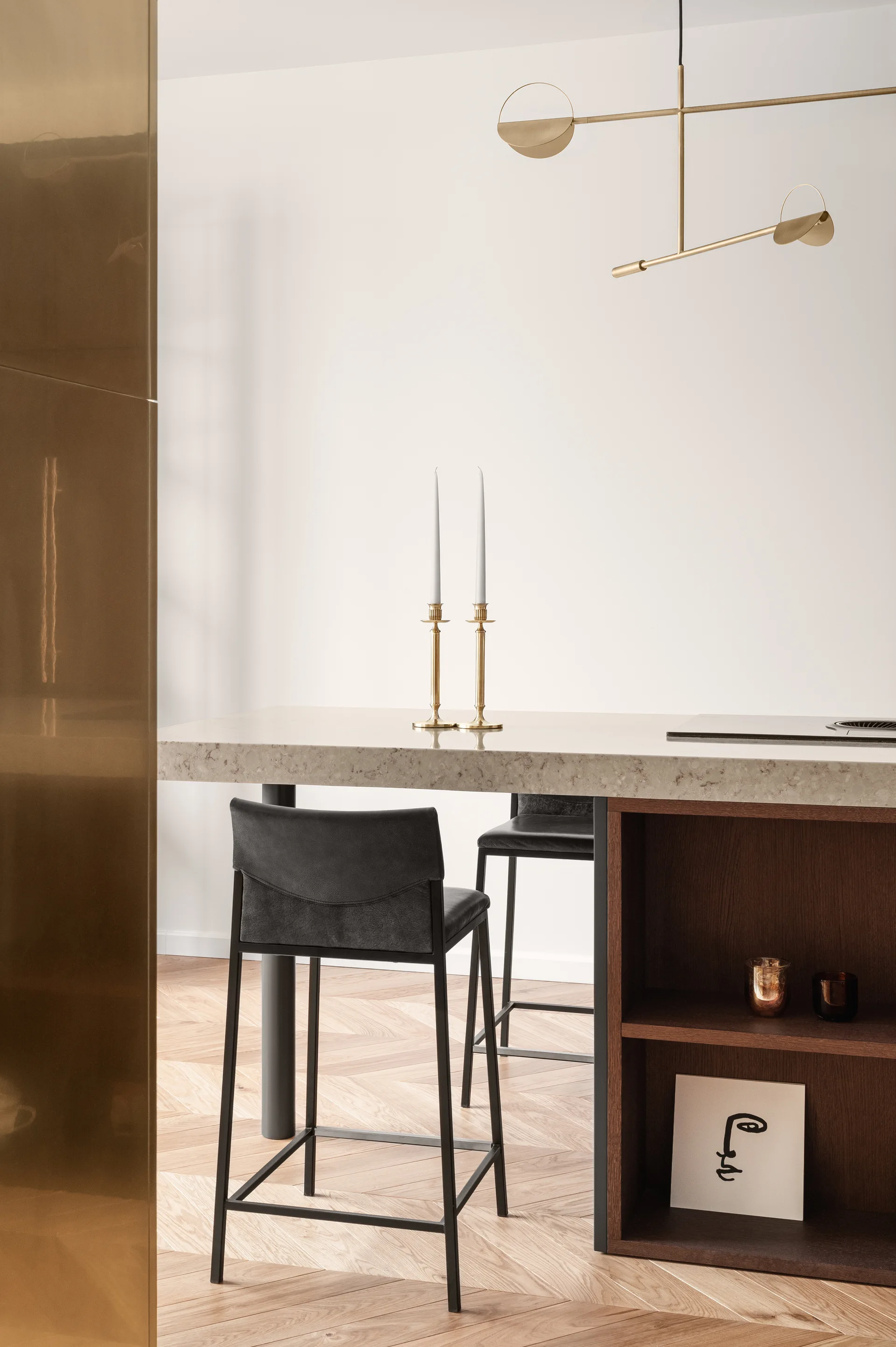 A light stone kitchen island features dark wooden shelves and black bar stools on herringbone parquet flooring beneath a brass pendant light.