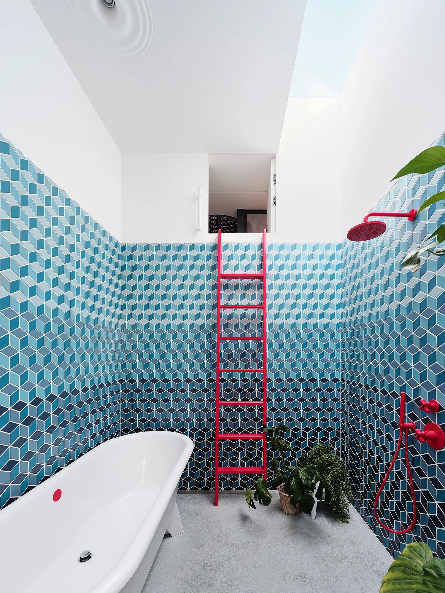A modern bathroom features blue geometric cube wall tiles, a white freestanding tub, a bright pink ladder, and matching pink shower fixtures.