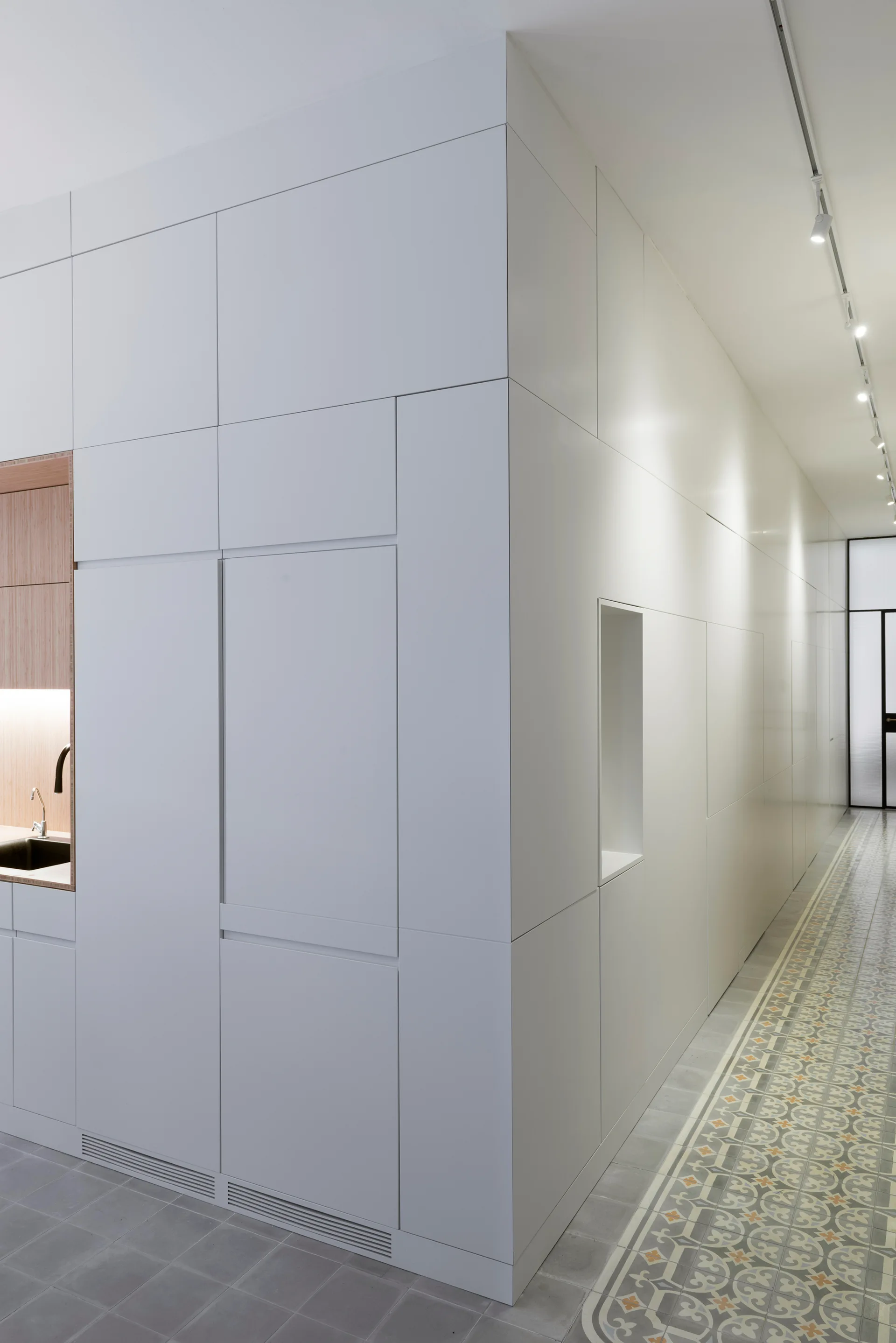 White floor-to-ceiling integrated cabinets with an inset wood-lined sink niche stand next to a hallway with patterned floor tiles.