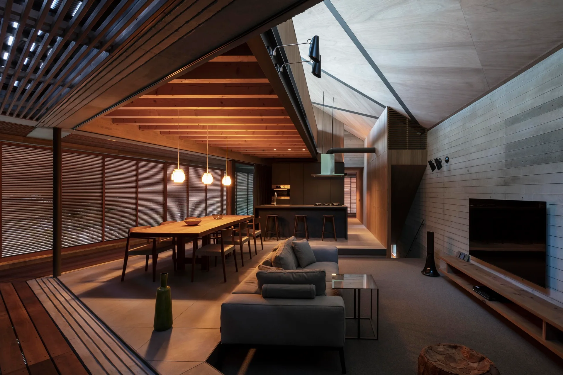 An open-plan living room features a gray upholstered sofa, a long wooden dining table under exposed timber beams, and a board-formed concrete wall.