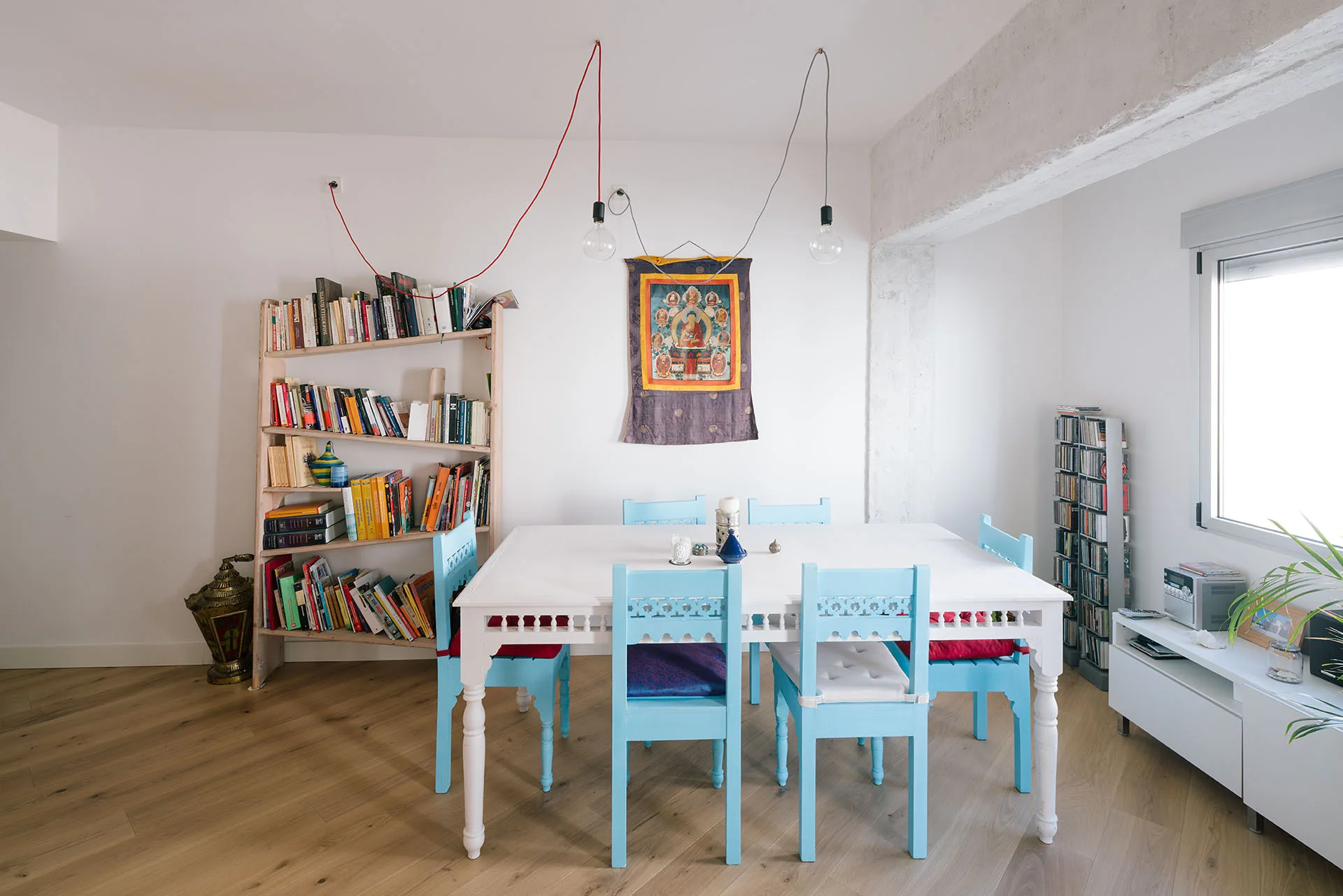 A bright dining room features a white table with light blue chairs, a leaning wooden bookshelf, and exposed ceiling beams on light hardwood floors.