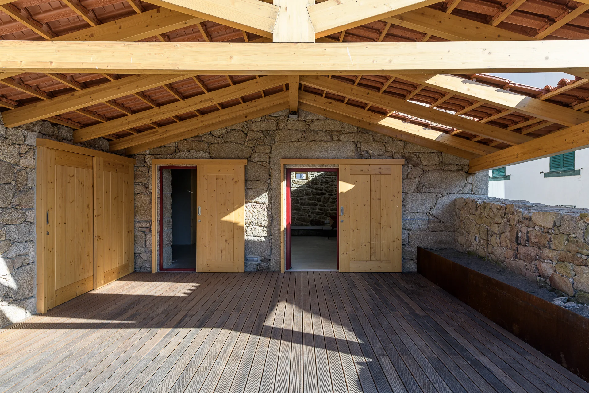 A covered outdoor patio with natural wood decking, rustic stone walls, sliding wooden doors with red trim, and an exposed timber truss roof.