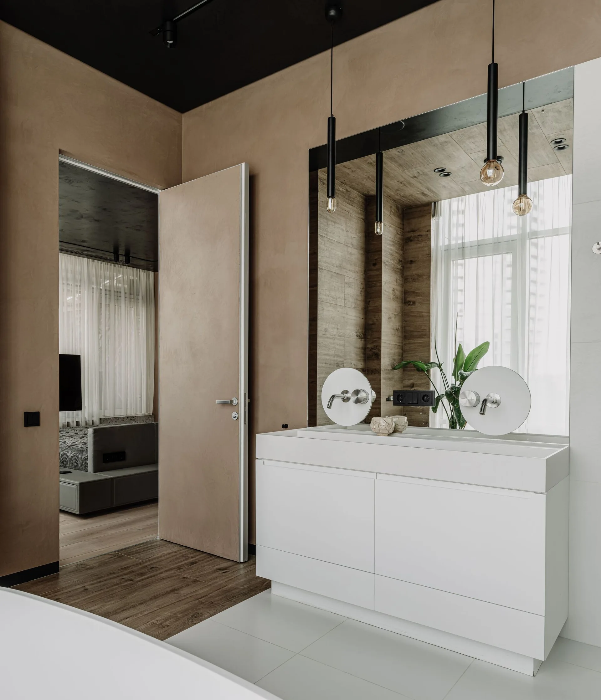 A modern bathroom features a white double vanity, round wall-mounted faucets, a large mirror, and slim black pendant lights against beige plaster walls.
