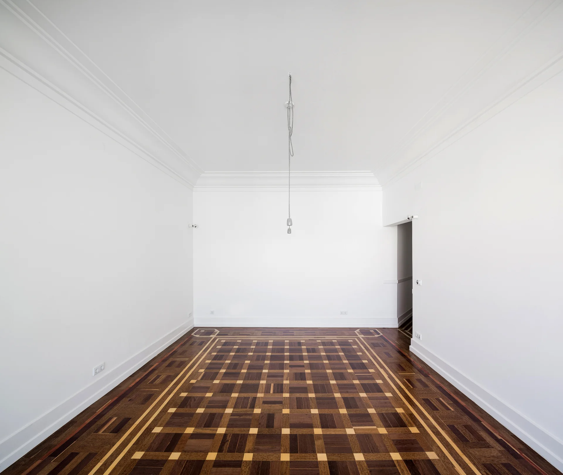 An empty room features bright white walls with decorative crown molding and an intricate geometric parquet wooden floor with a single hanging pendant cord.