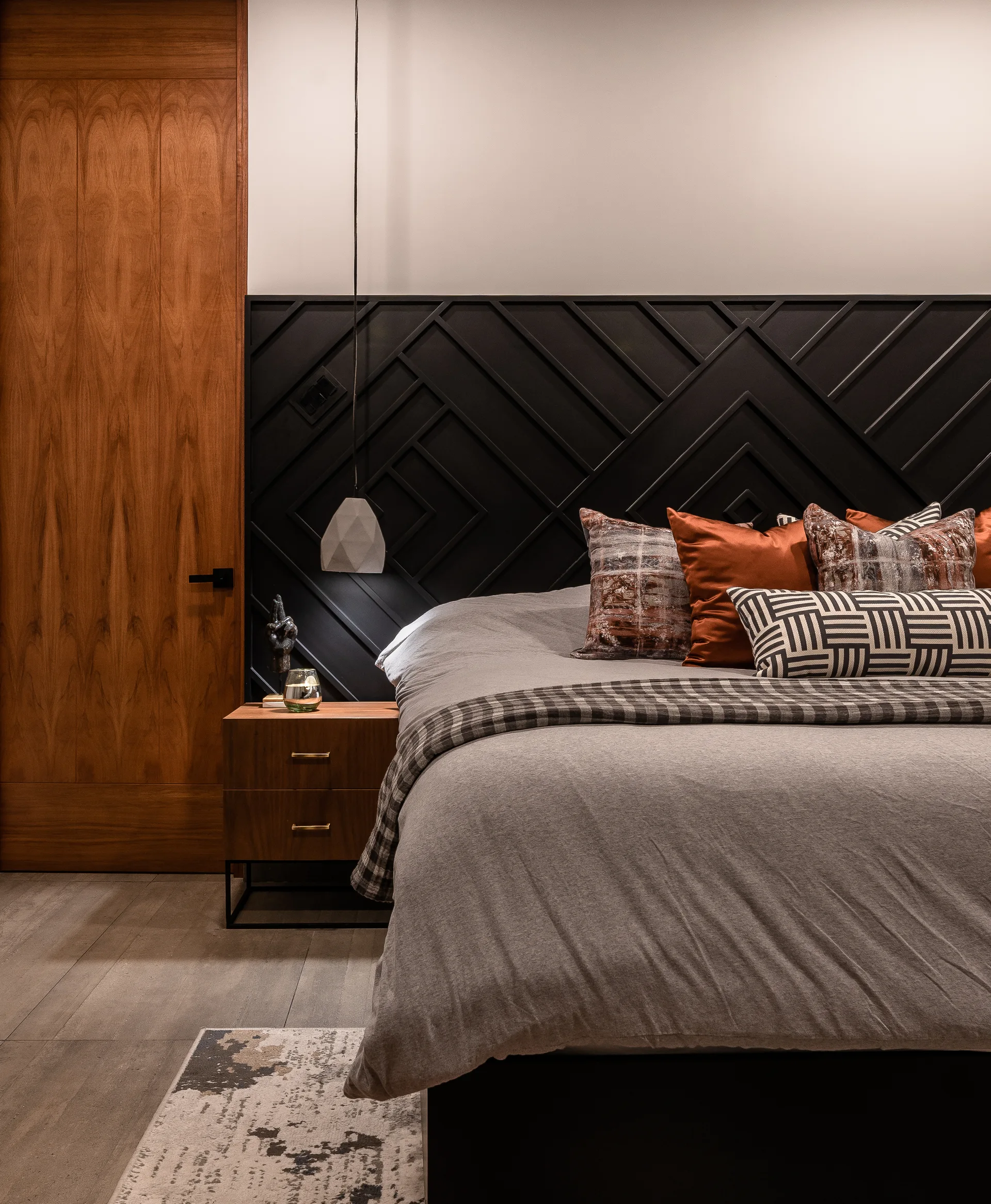 A modern bedroom features grey bedding on a bed set against a black geometric patterned wood headboard wall next to a warm wood nightstand.
