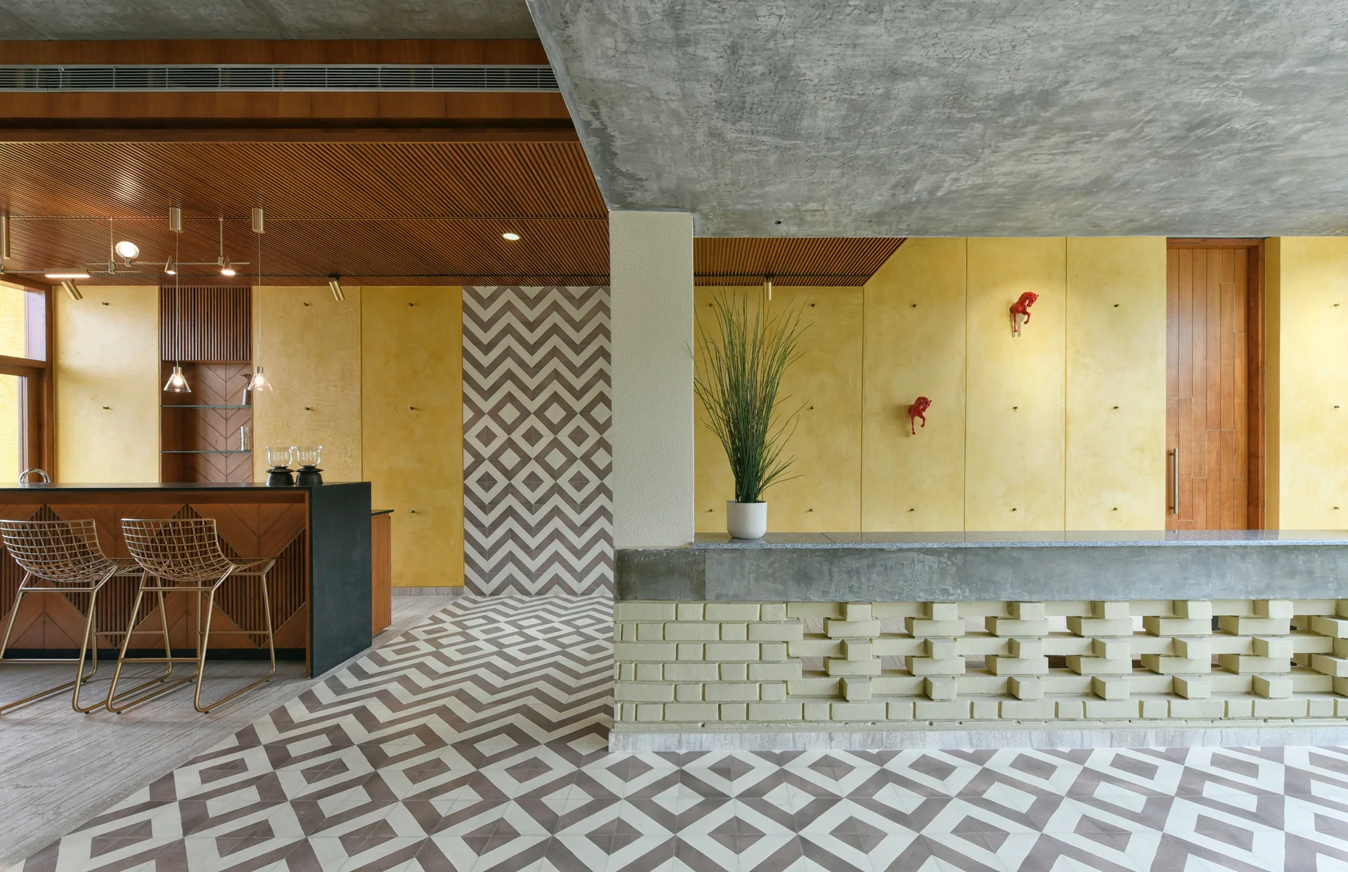 An interior bar area featuring yellow plaster walls, gold-toned wire bar stools, and bold geometric tile patterns on the floor.