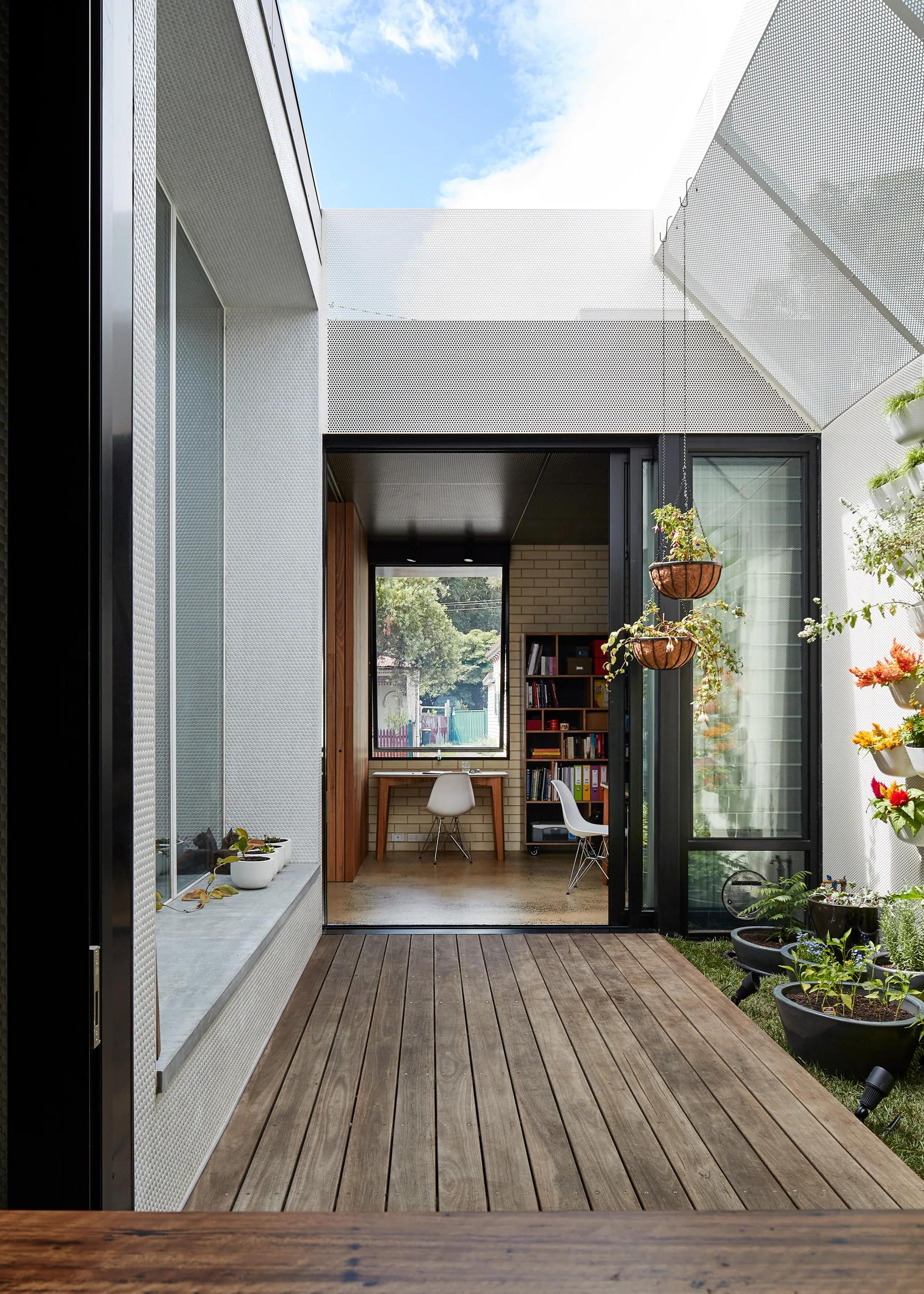 A wooden patio deck with hanging plant baskets leads through sliding glass doors to a home office featuring a desk and bookshelves.