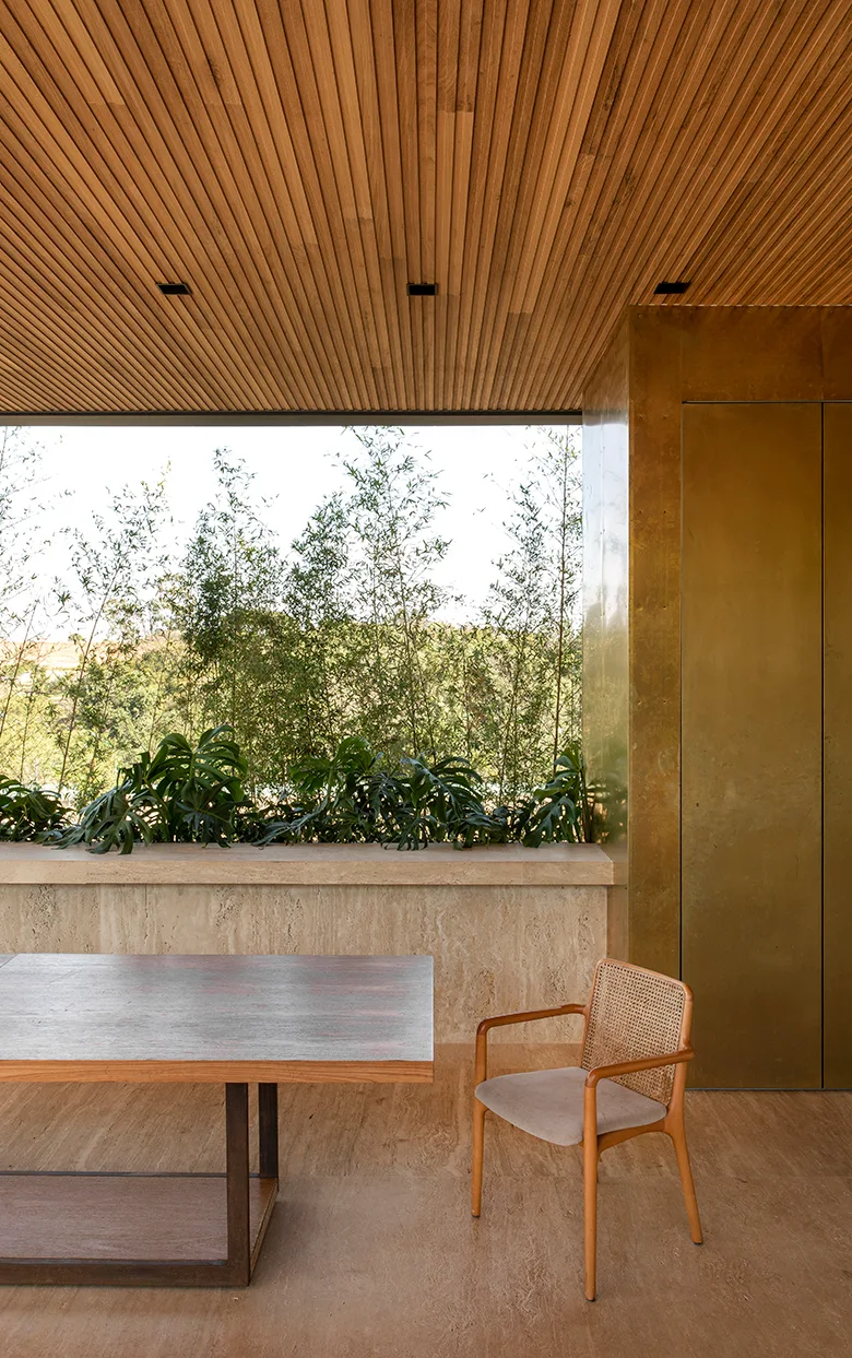 A modern dining area showcases a slatted wood ceiling, a gold metallic wall panel, and a cane-back chair near a stone ledge and window.