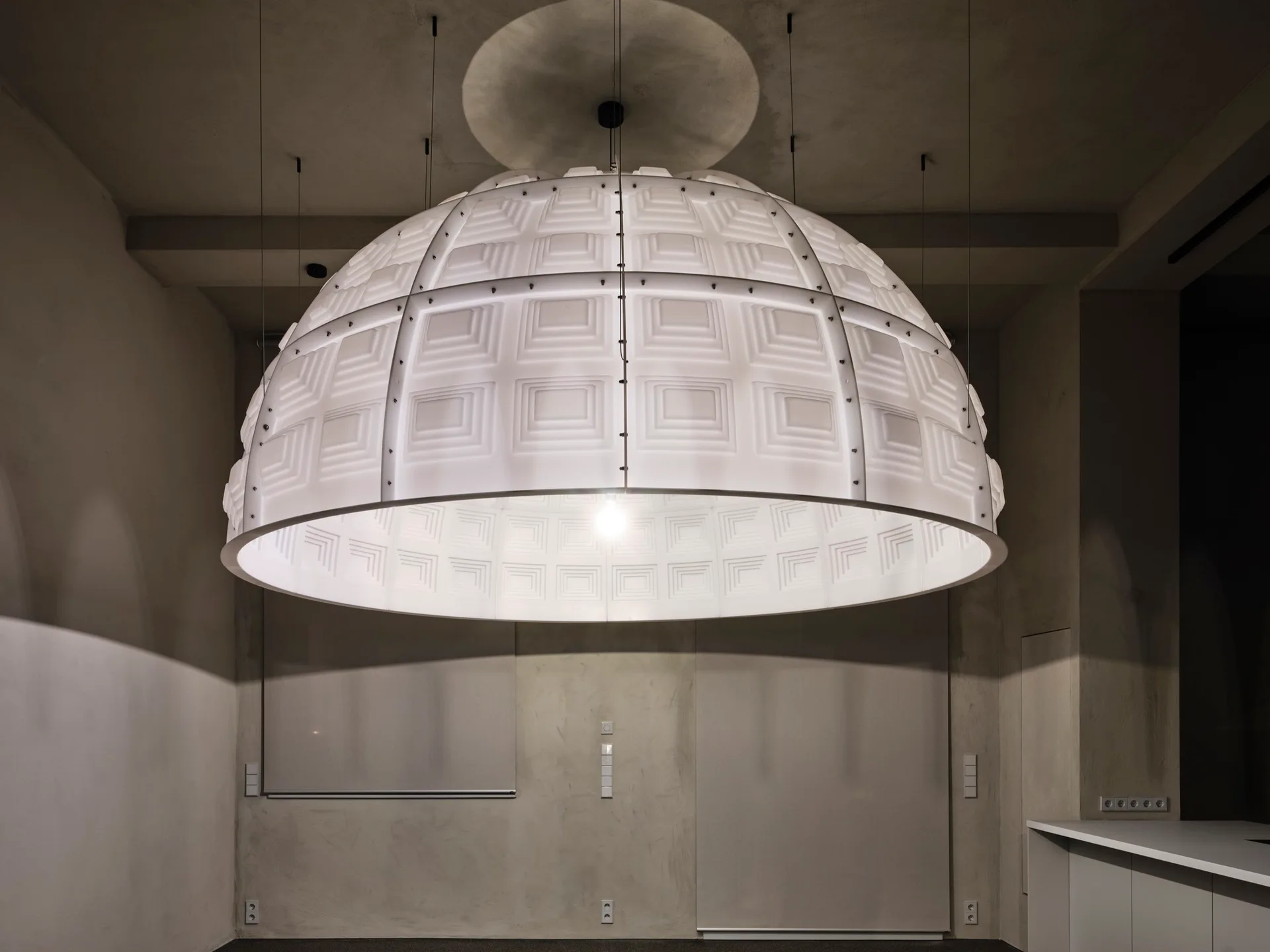 A large, white dome-shaped pendant light with geometric square patterns hangs from the ceiling in a minimalist grey concrete room.