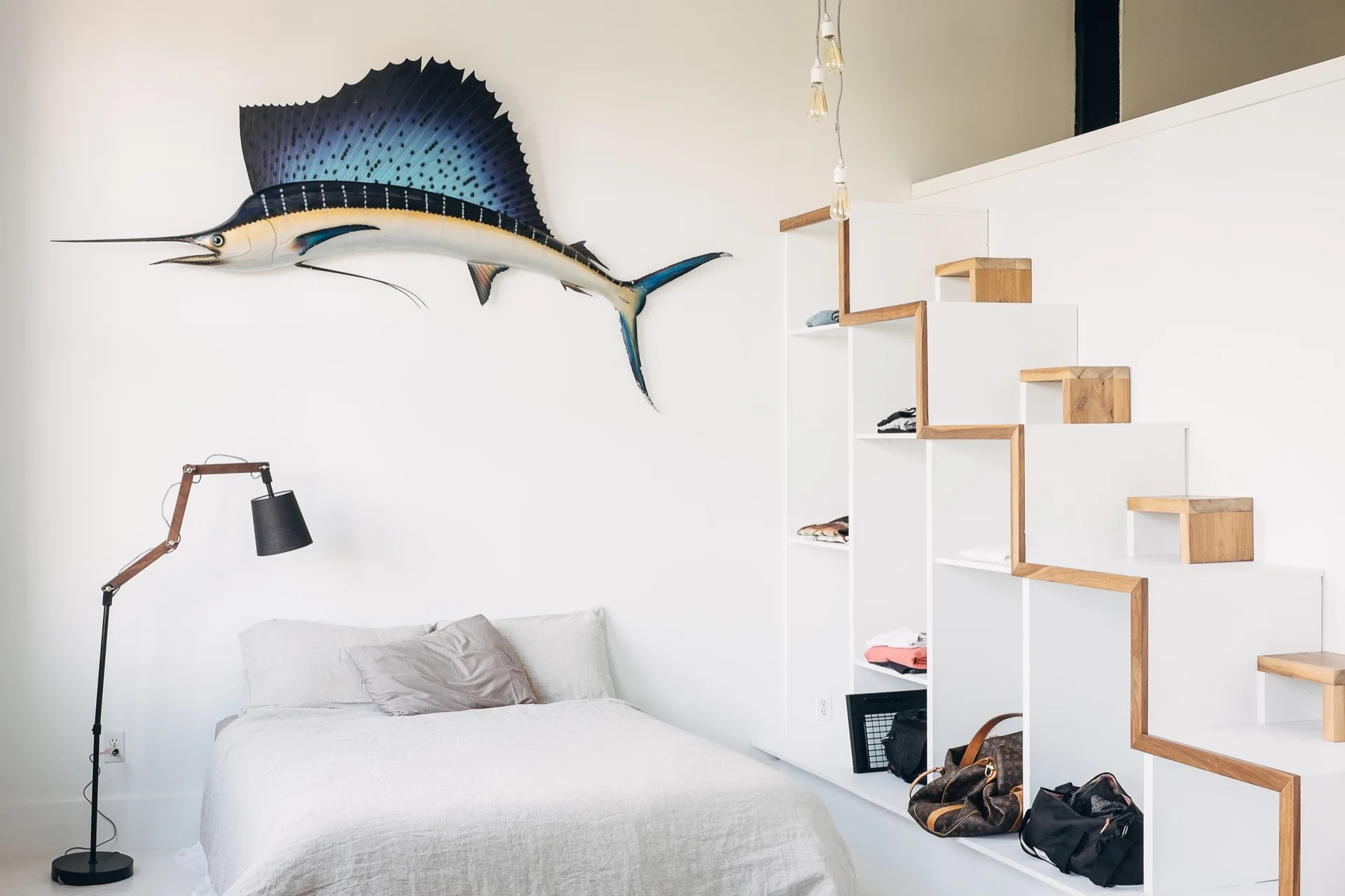 A minimalist bedroom displays a low bed with grey bedding, a blue sailfish wall mount, and a white integrated staircase shelving unit with wood accents.