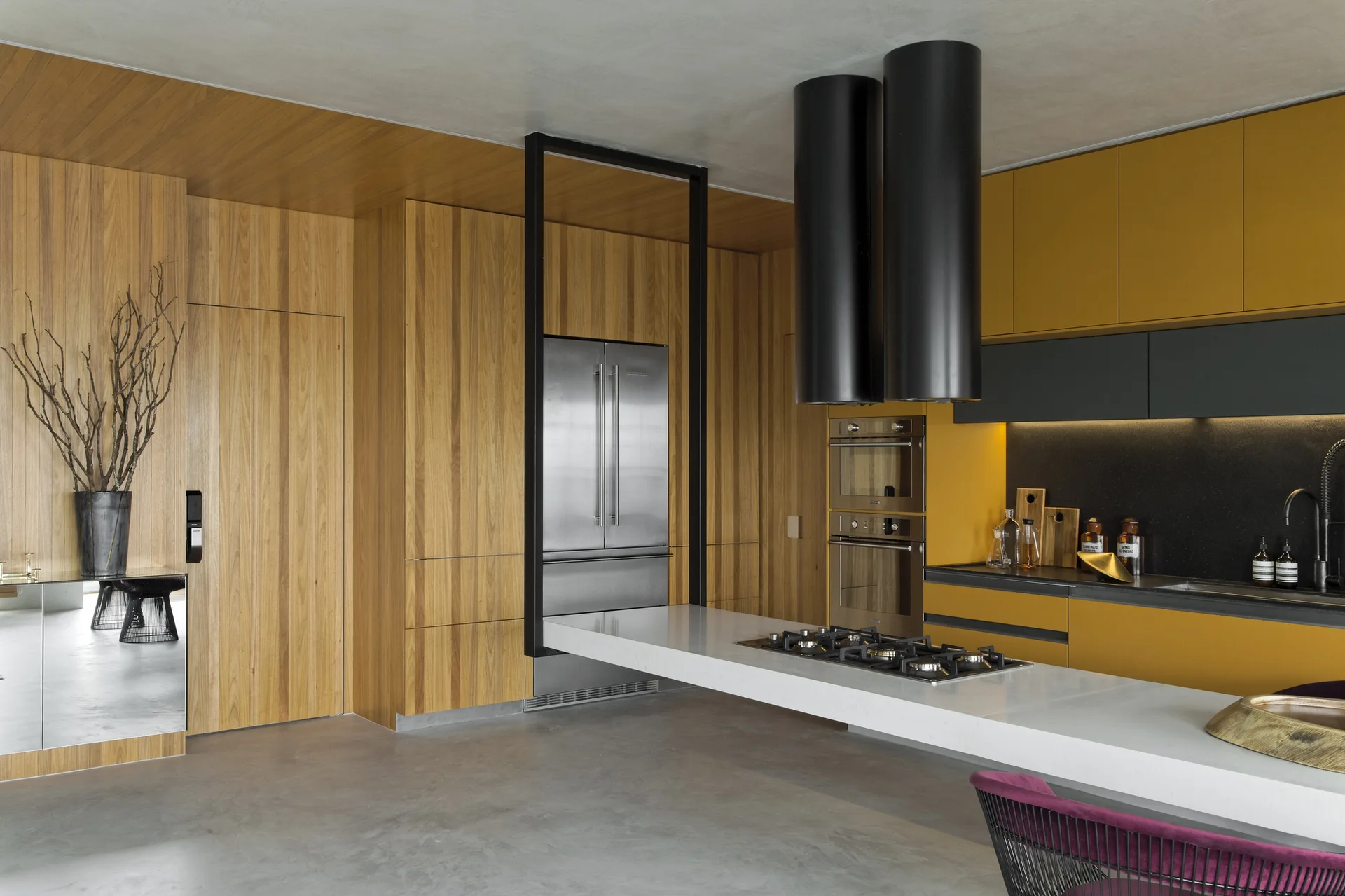 A modern kitchen featuring mustard yellow cabinetry, warm wood wall panels, a long white island, and twin black cylindrical range hoods.