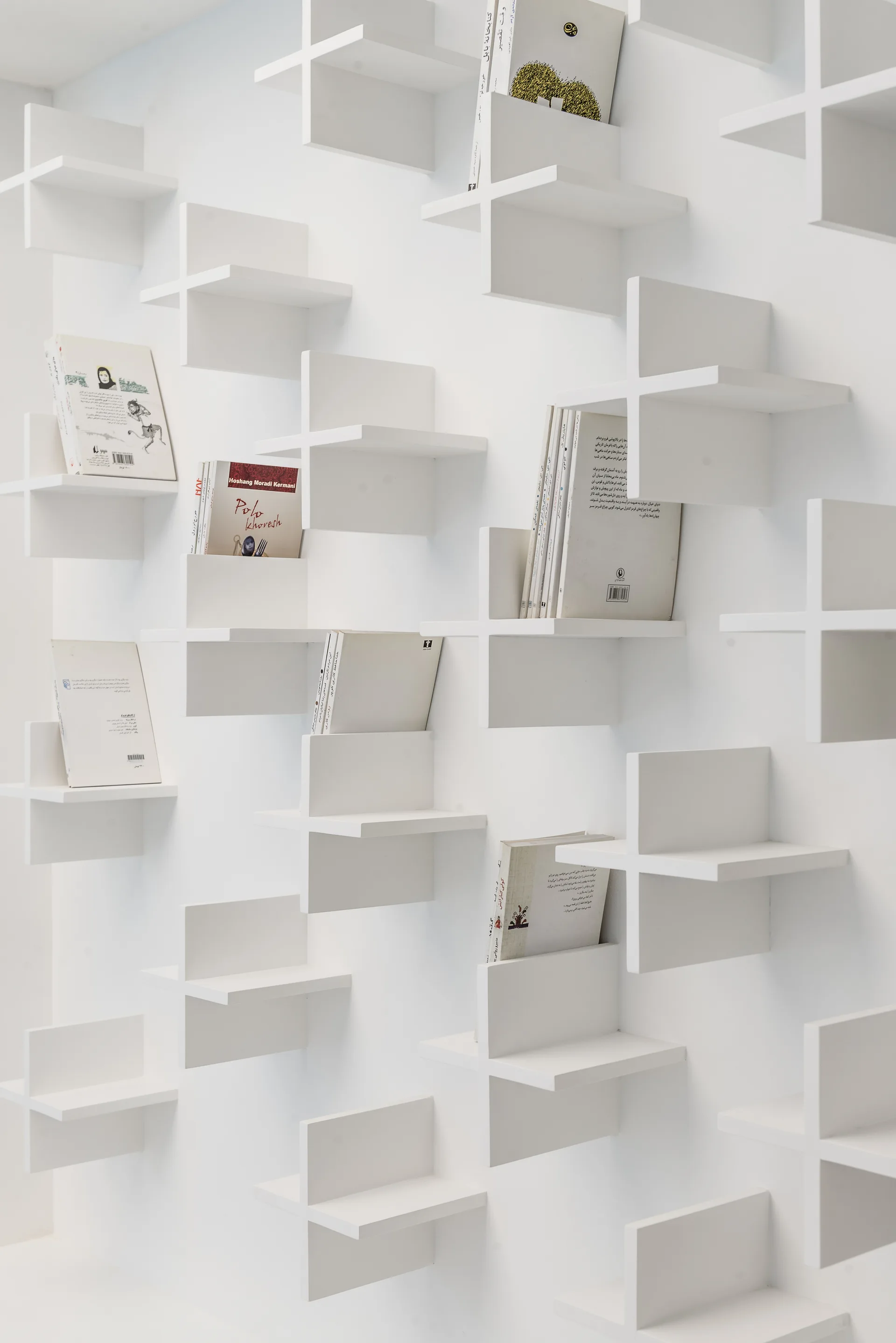 Several minimalist white cross-shaped shelves are mounted in a staggered geometric pattern on a flat white wall holding a few books.