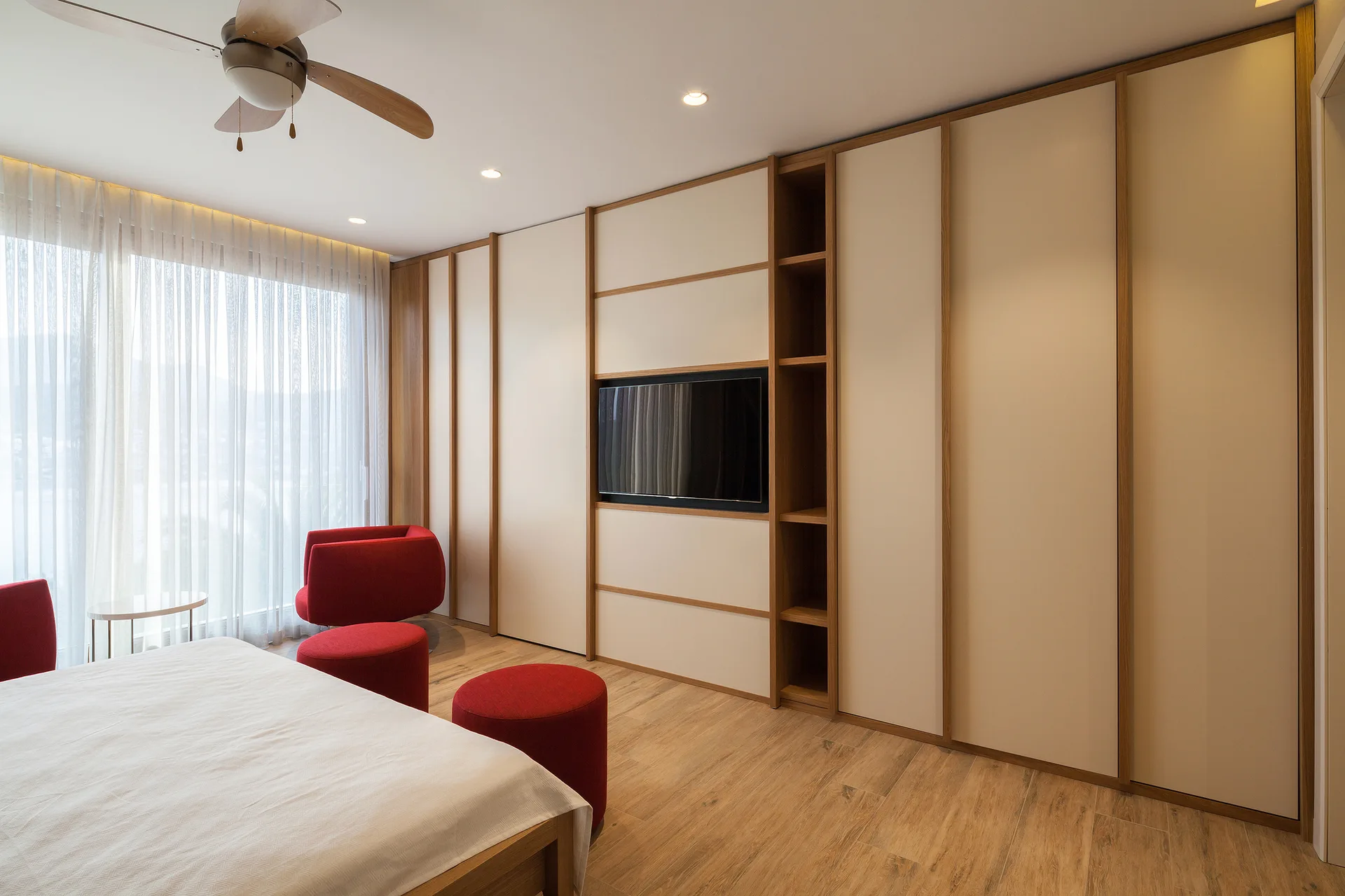 A modern bedroom features a large beige and wood built-in wardrobe with an integrated television alongside a bed and bright red accent chairs.